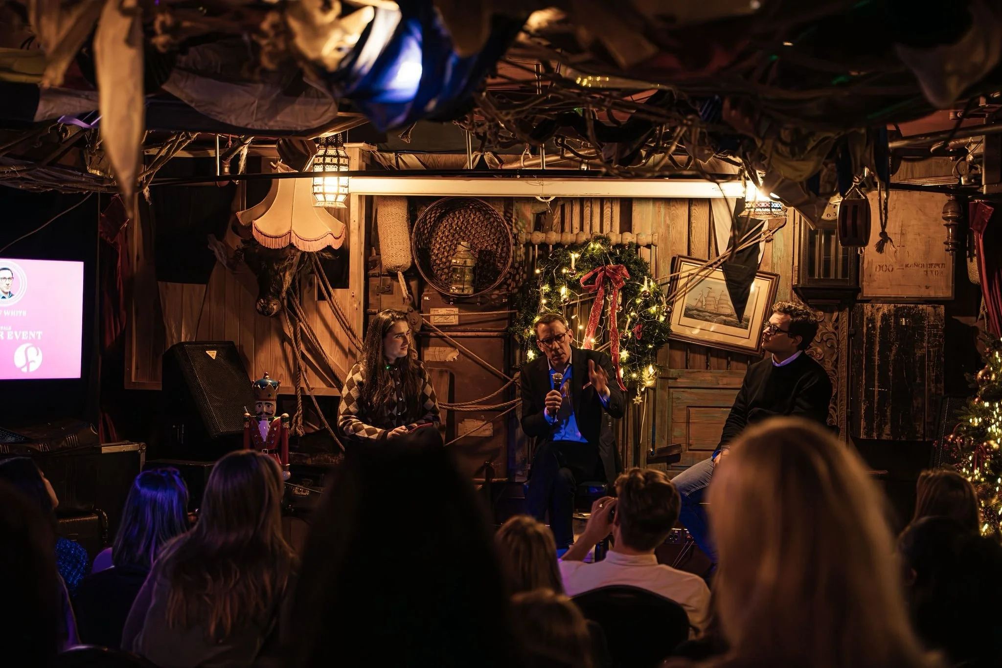 A man speaking into a microphone during a panel discussion on a small stage decorated with a Christmas wreath and holiday decorations, with three people seated on the stage and an audience watching in a cozy, rustic venue.