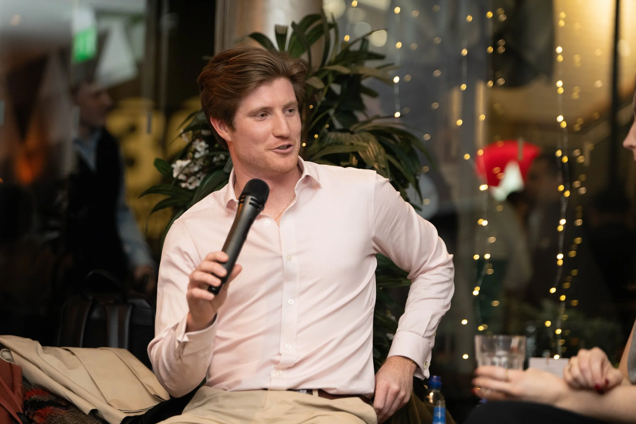 Man with brown hair in a pink shirt holding a microphone, engaging in conversation with a woman holding a glass, sitting in a cozy, decorated indoor setting with string lights and plants.