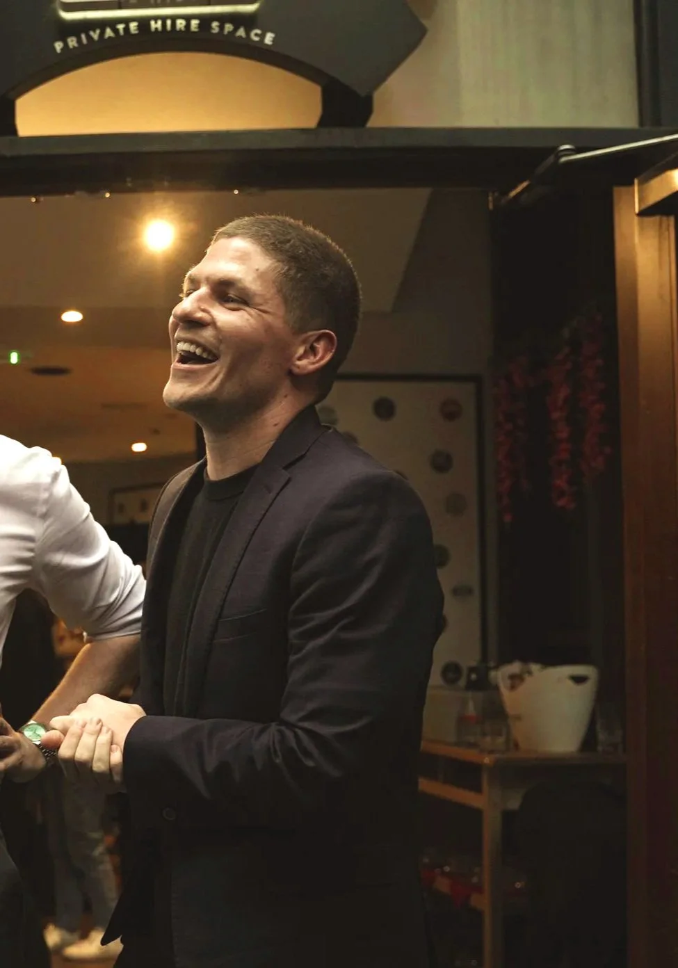 A man laughing and smiling, dressed in a black suit, in a dimly lit indoor setting with a sign that reads 'PRIVATE HIRE SPACE' in the background.