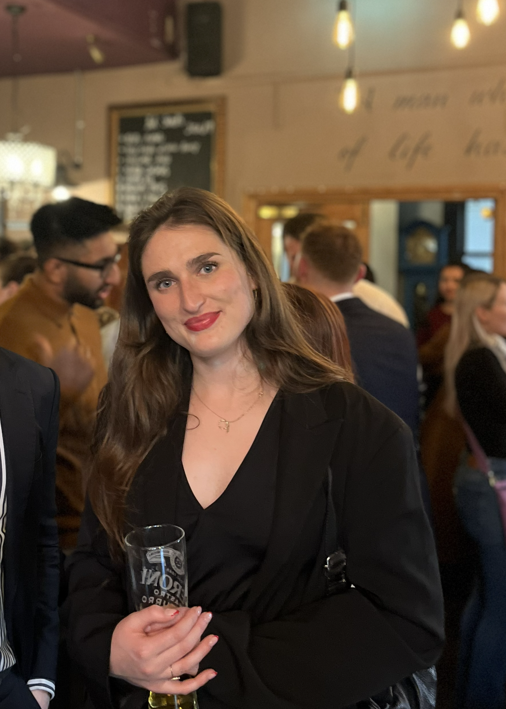 A woman with long brown hair and red lipstick holding a clear glass of beer in a crowded restaurant or bar with warm lighting.
