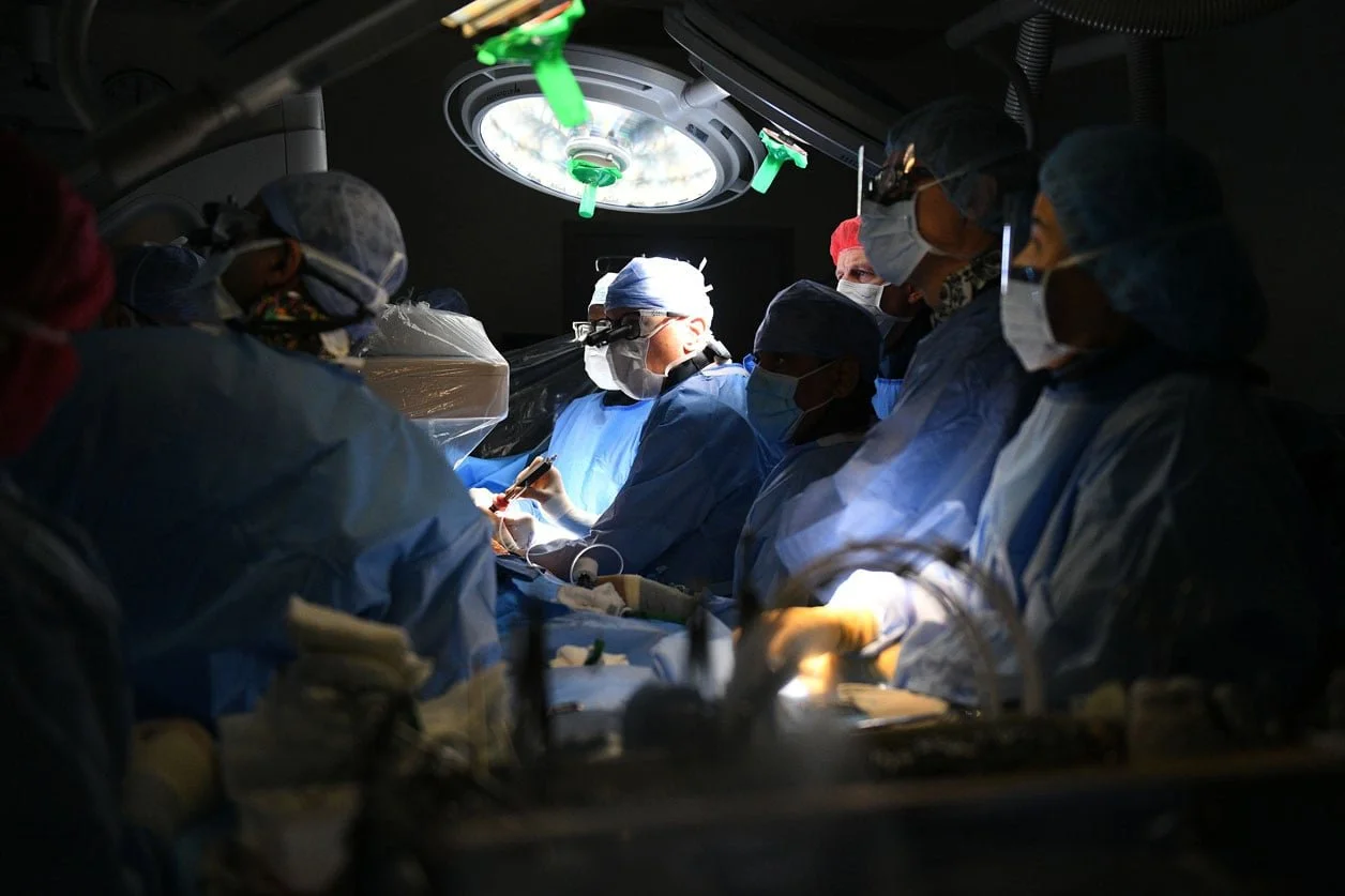 Surgeons performing a surgical operation in an operating room, wearing surgical masks, caps, and gowns, with a bright overhead surgical light illuminating the procedure.