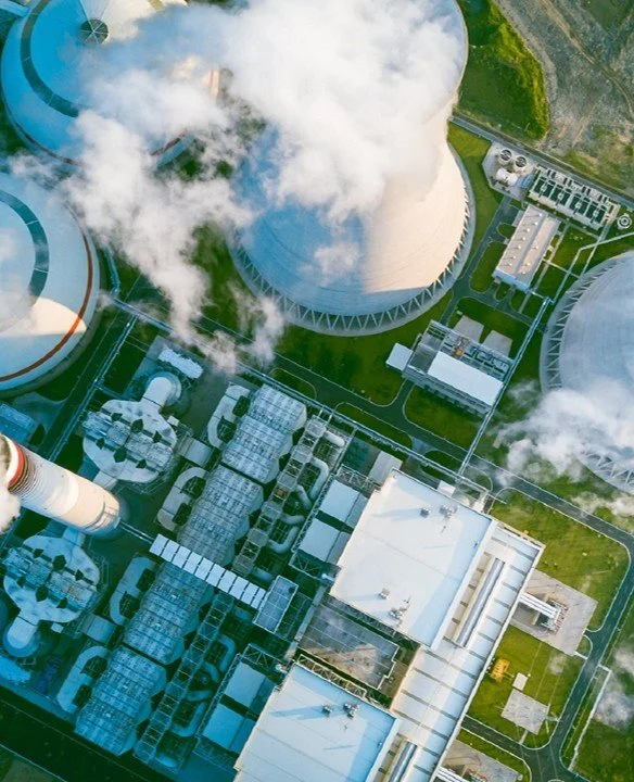 Aerial view of a nuclear power plant with cooling towers releasing steam, surrounded by industrial structures and greenery.