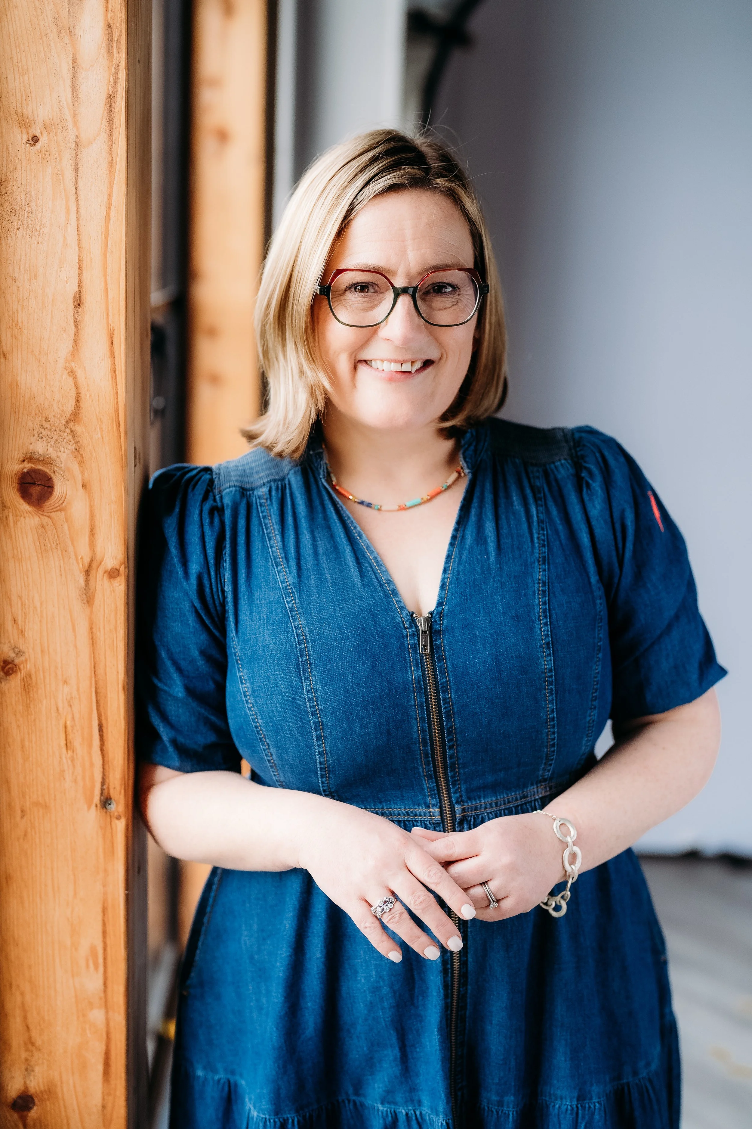 A woman with glasses and shoulder-length blonde hair smiling in a denim dress, standing indoors beside a wooden wall.