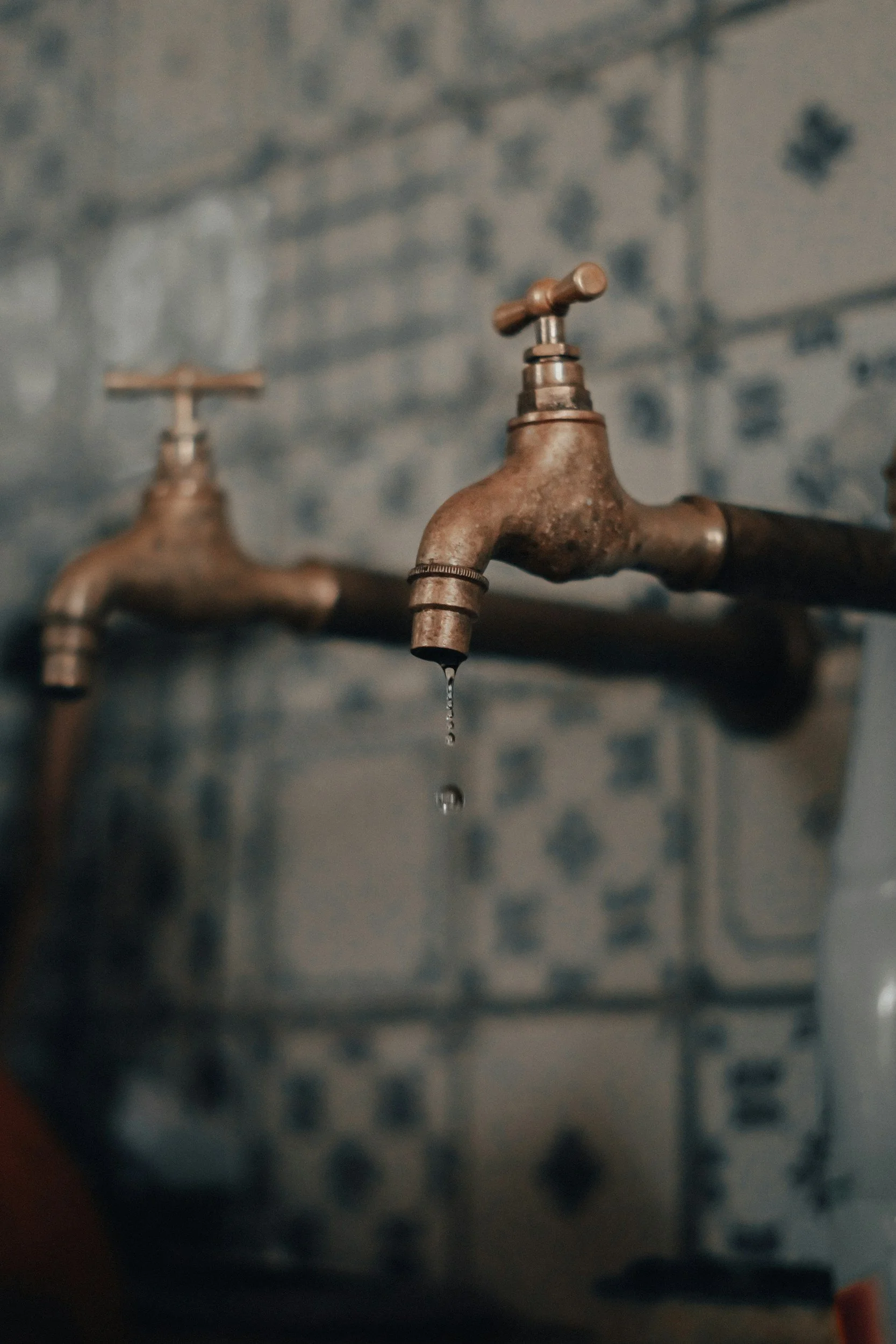 An old, rusty faucet with water droplet dripping from it, set against a blue and white tiled wall.