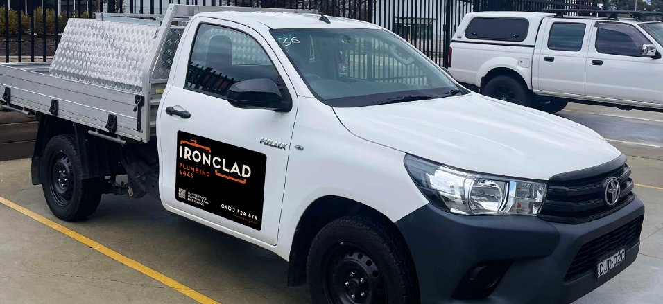 White flatbed truck with a black and orange Ironclad Plumbing & Gas logo on the door, parked in a lot with other vehicles visible in the background.