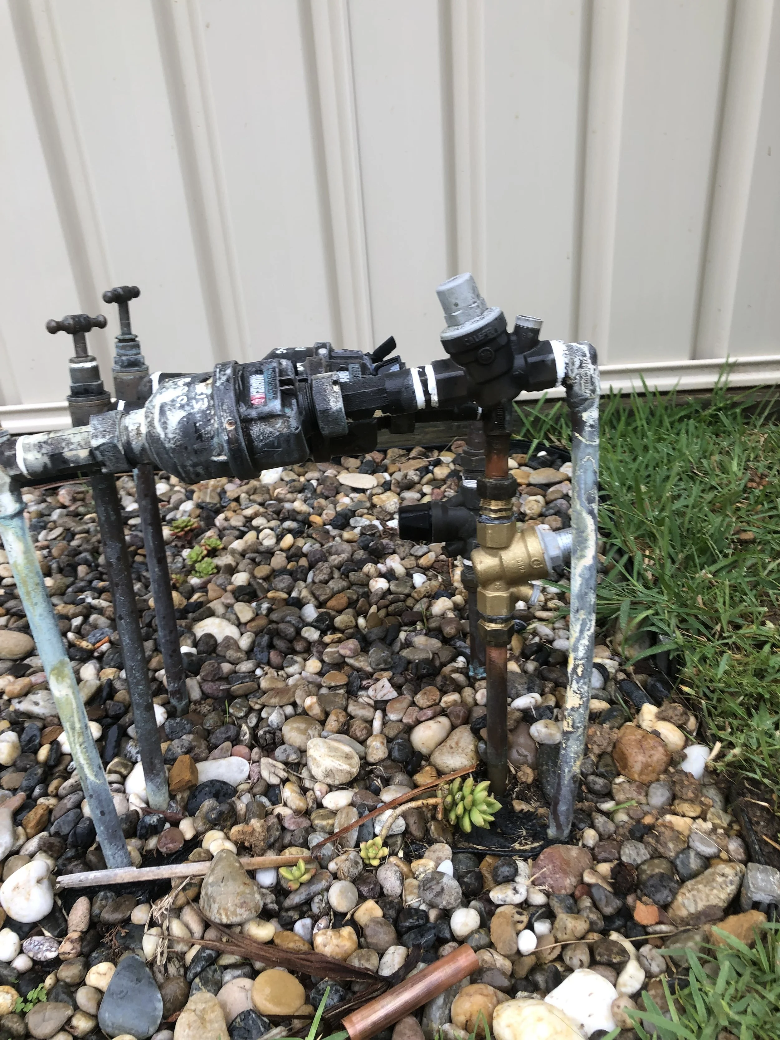 Outdoor sprinkler system with pipes and valves installed on a bed of multicolored rocks and nearby green grass, with a white siding wall in the background.