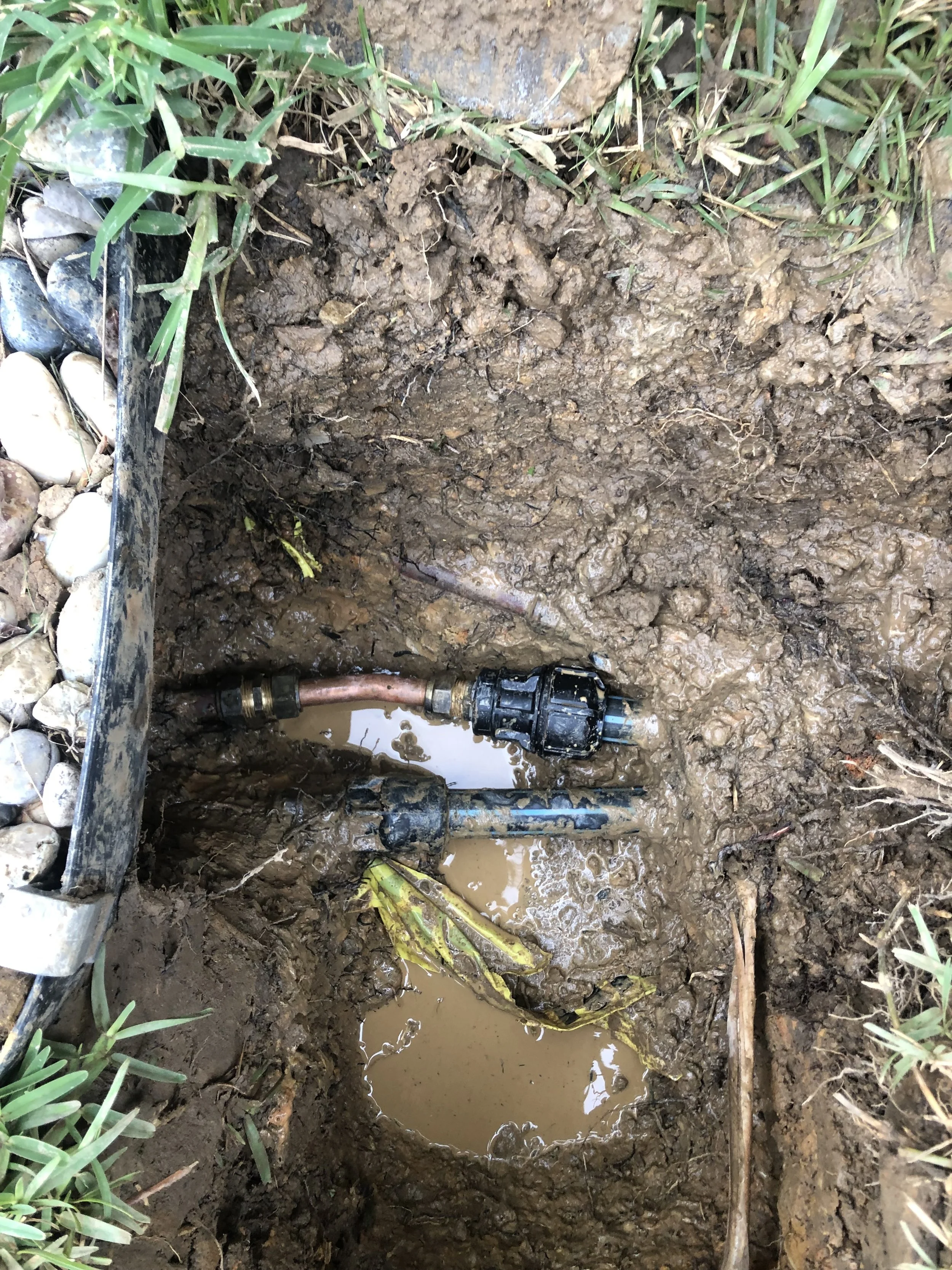 A close-up of a water pipe connection in a muddy hole, with muddy water pooled around it, surrounded by grass and small rocks.