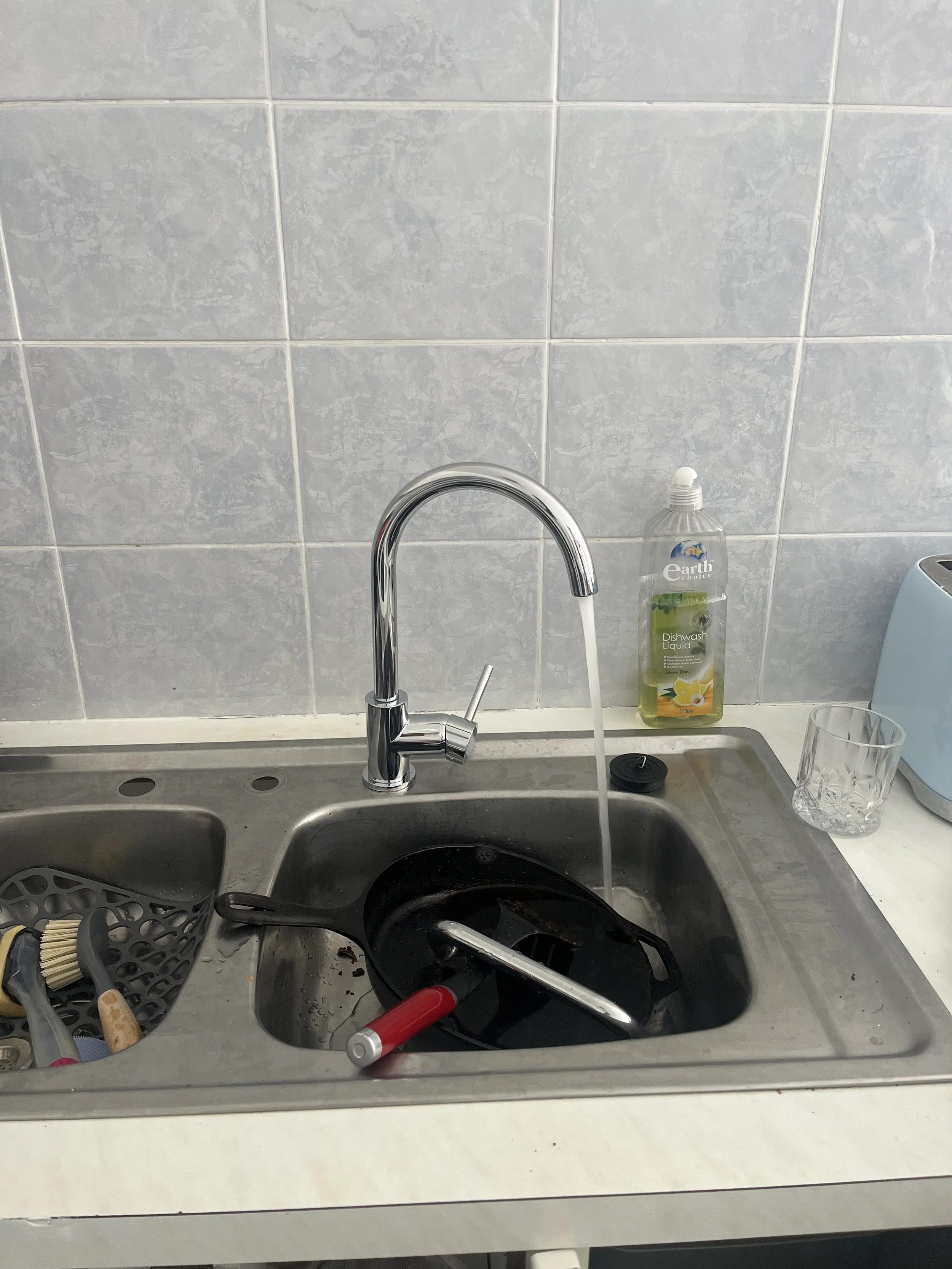 Kitchen sink with running water, a dish scrubber, black cooking pan, red-handled kitchen tool, dish soap bottle, glass, and toaster on silver countertop.