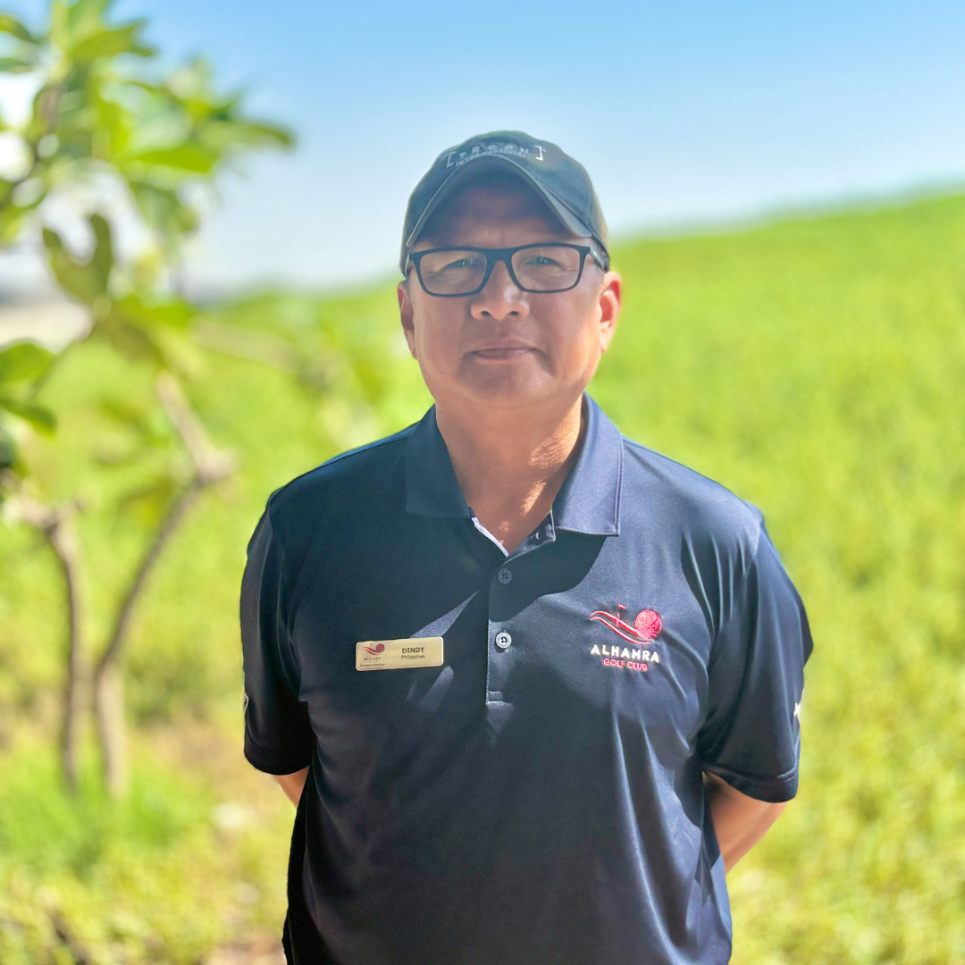 A man wearing glasses, a black cap, and a navy blue polo shirt with a logo that says 'Alhambra Golf Club' stands outdoors on a sunny day with green fields in the background.