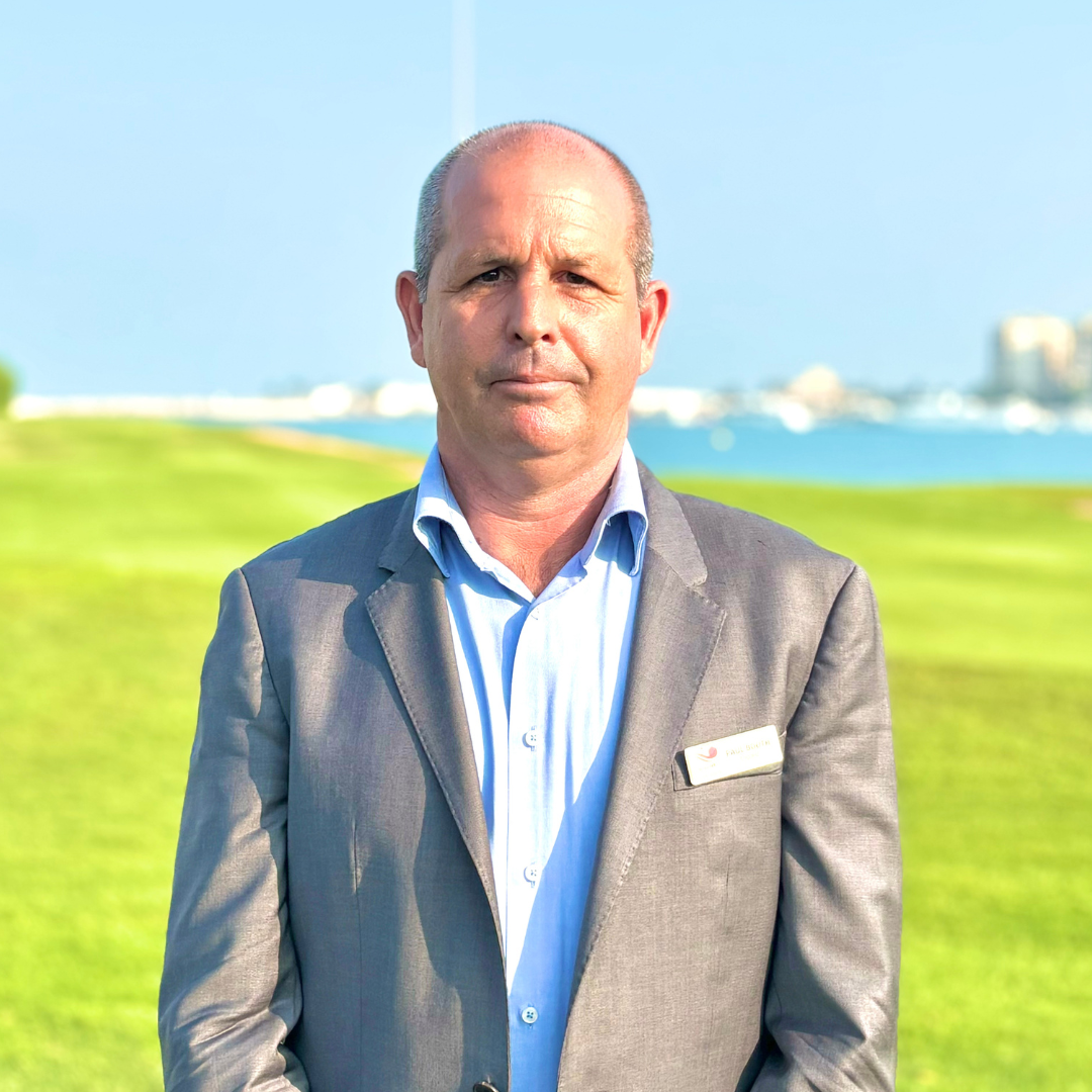 A man in a gray suit standing outdoors on a grassy area with water and buildings in the background.