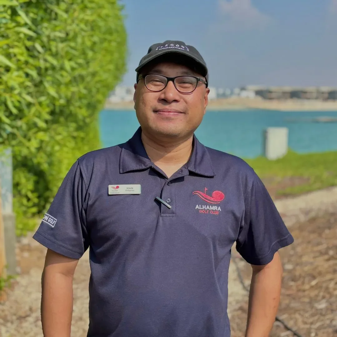 A man smiling outdoors wearing glasses, a cap, and a navy blue polo shirt with the Alhamra Golf Club logo.