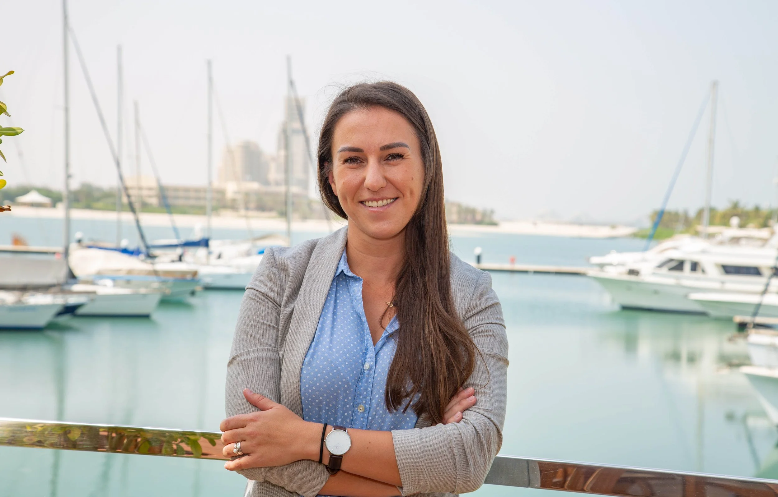 A woman with long brown hair wearing a grey blazer and a blue polka dot shirt, smiling with arms crossed at a marina with sailboats and yachts in the background.
