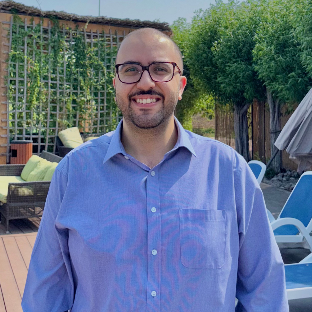 A smiling man with glasses and a beard wearing a light blue button-up shirt outdoors in a backyard with patio furniture, trees, and a wooden fence.