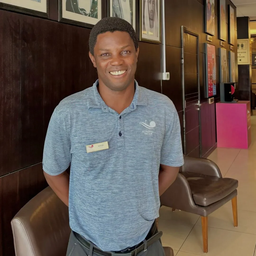 Smiling man wearing a gray polo shirt with a name tag, standing in a lobby with framed artwork on the wall.