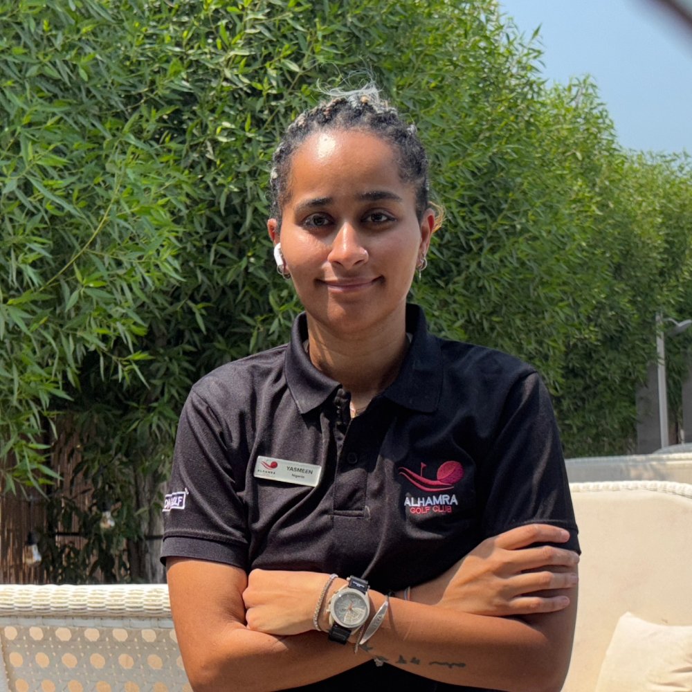 A woman with braided hair, wearing a black polo shirt with a name tag and ALHAMRA GOLF CLUB logo, standing outdoors with green foliage in the background.