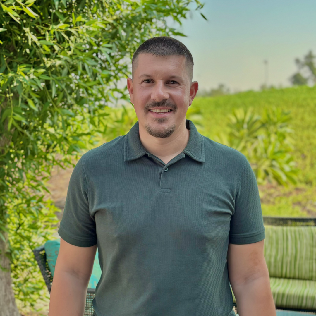 A man with short dark hair, a goatee, and a mustache smiling outdoors in front of green foliage and a field.