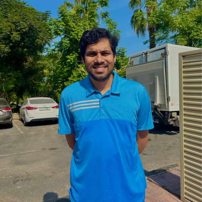 Man in a blue polo shirt standing outdoors with trees and parked cars in the background.