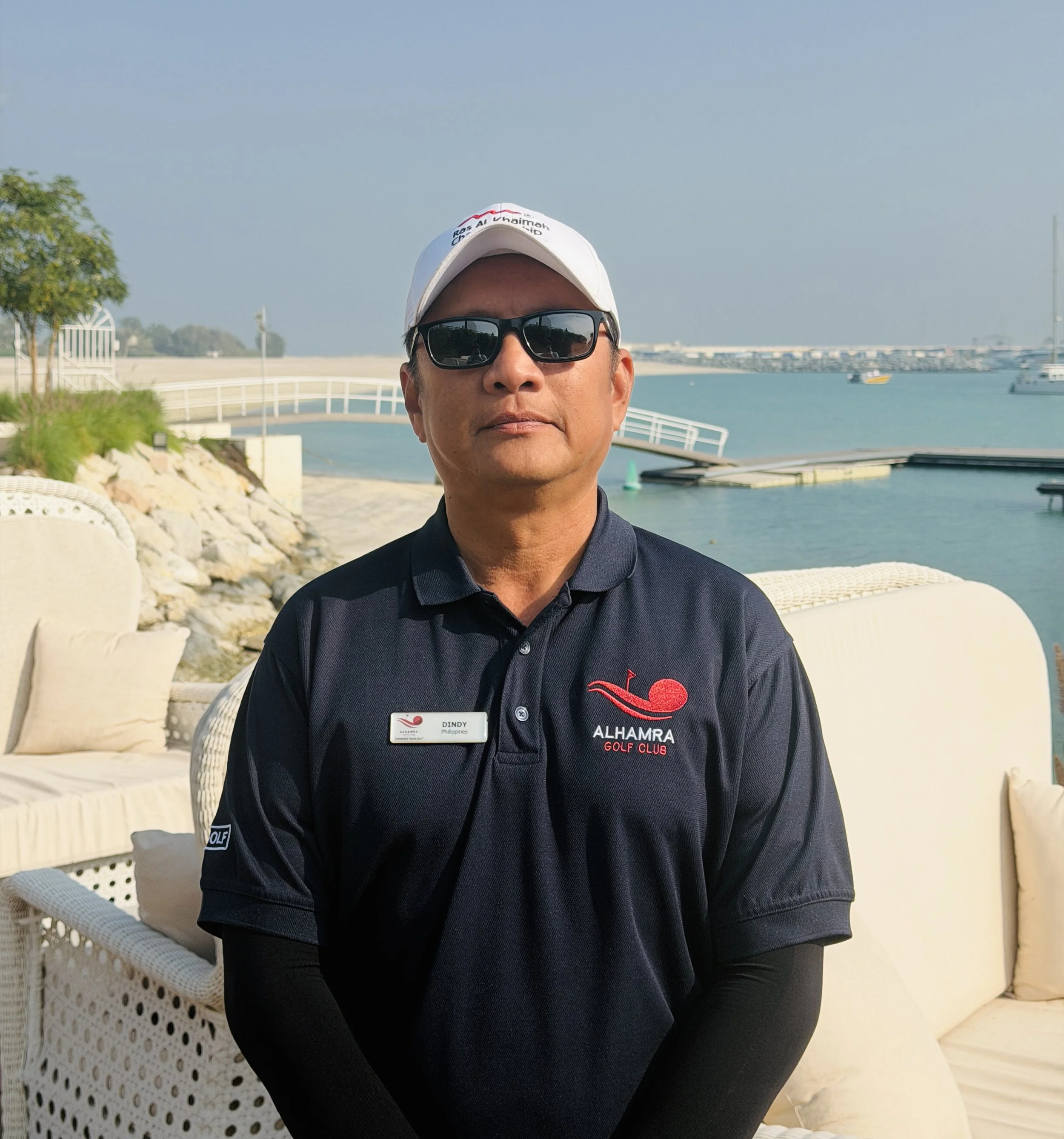 A man wearing glasses, a black cap, and a navy blue polo shirt with a logo that says 'Alhambra Golf Club' stands outdoors on a sunny day with green fields in the background.