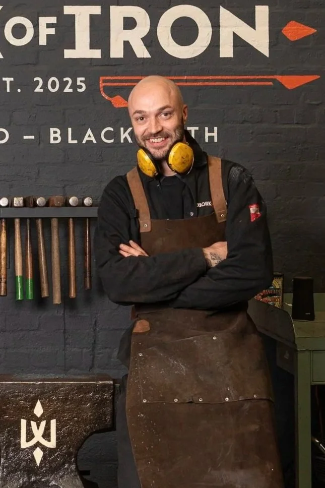 A man with tattoos on both arms, wearing a brown leather apron, stands in a blacksmith's workshop, with a forge and tools visible behind him.