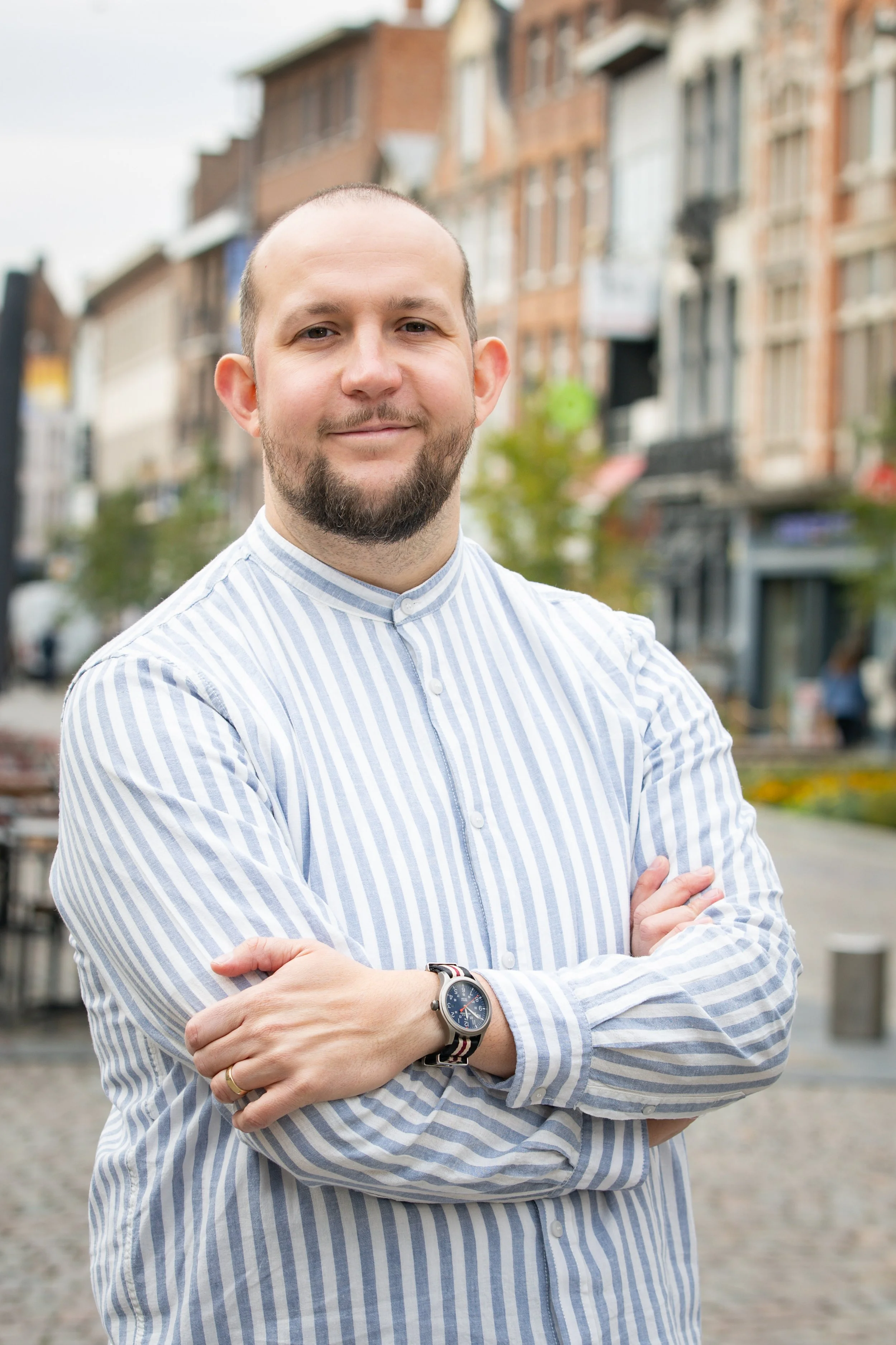 A man with a beard and short hair, wearing a striped button-up shirt and a watch, standing outdoors with his arms crossed on a street with buildings and trees in the background.