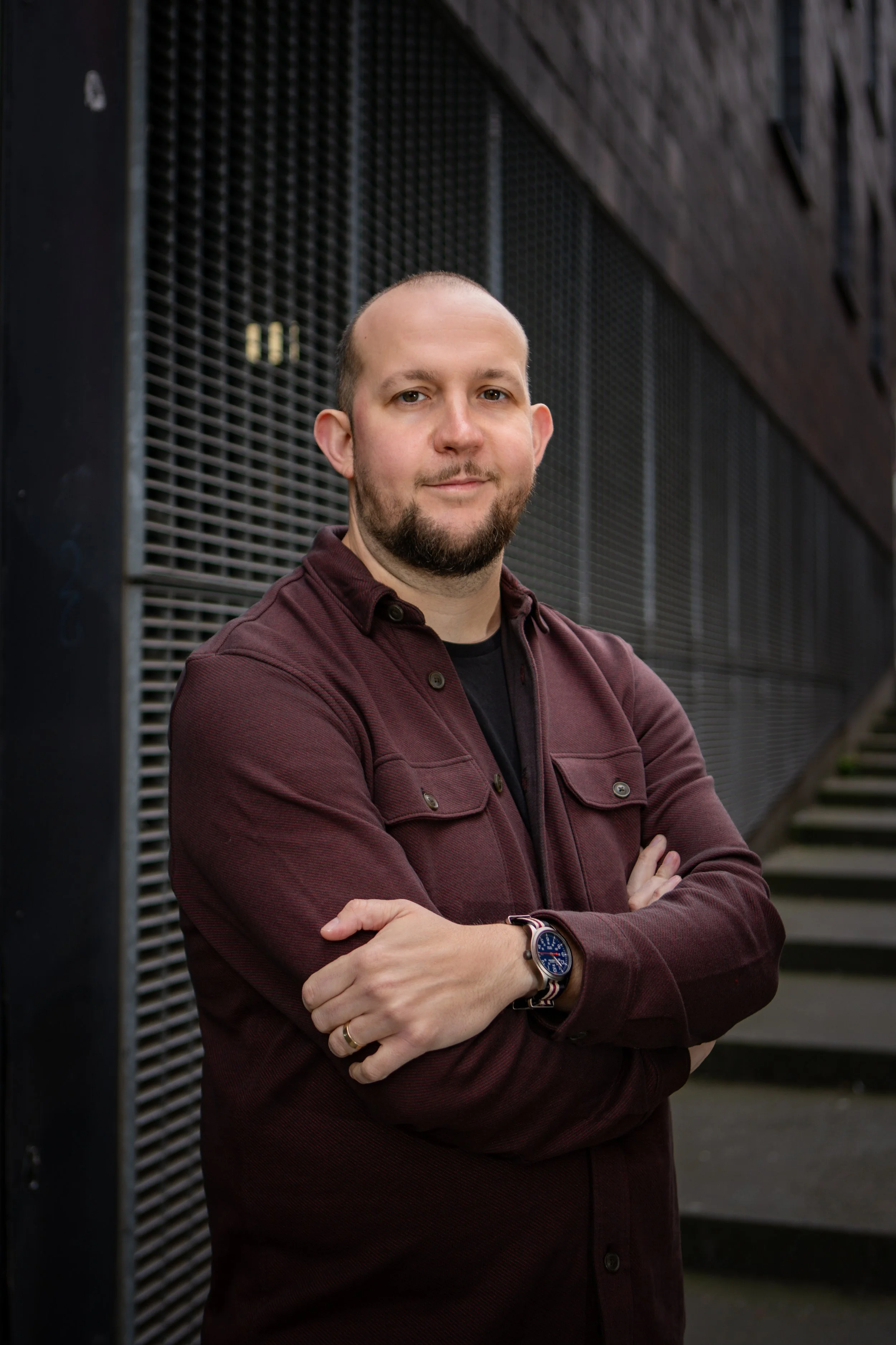 A man with a beard and short hair, wearing a maroon jacket and a watch, standing with arms crossed in front of a modern building with black metal panels and stairs.