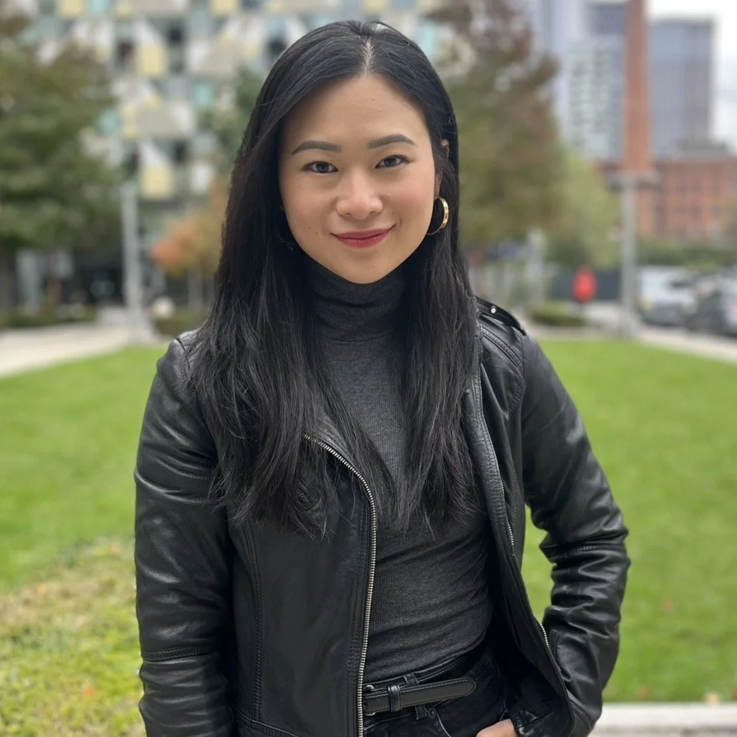 A woman with long black hair, wearing a dark turtleneck and leather jacket, standing outdoors in a park with green grass and trees, city buildings in the background.