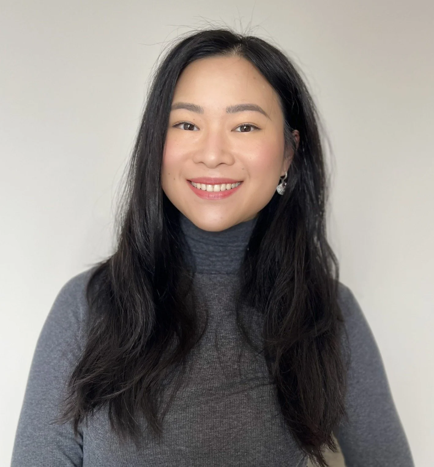 A young woman with long black hair, smiling, wearing a gray turtleneck sweater and earrings, standing against a plain light-colored background.