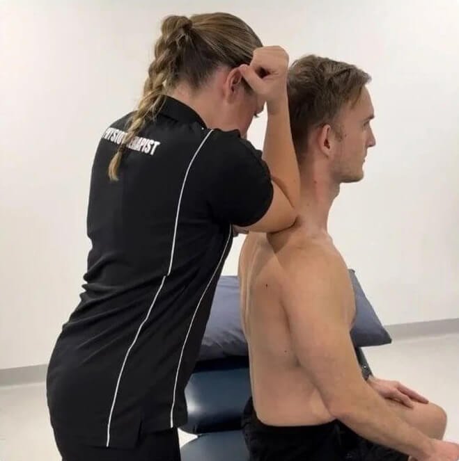 A man practicing a fitness exercise with a woman on a gray carpeted floor in a room with shelves. The man is holding the woman, who is seated, while she leans back with her arms extended.