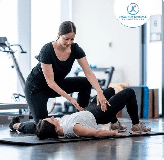 A woman exercises on a stability ball while a trainer assists her in a gym.