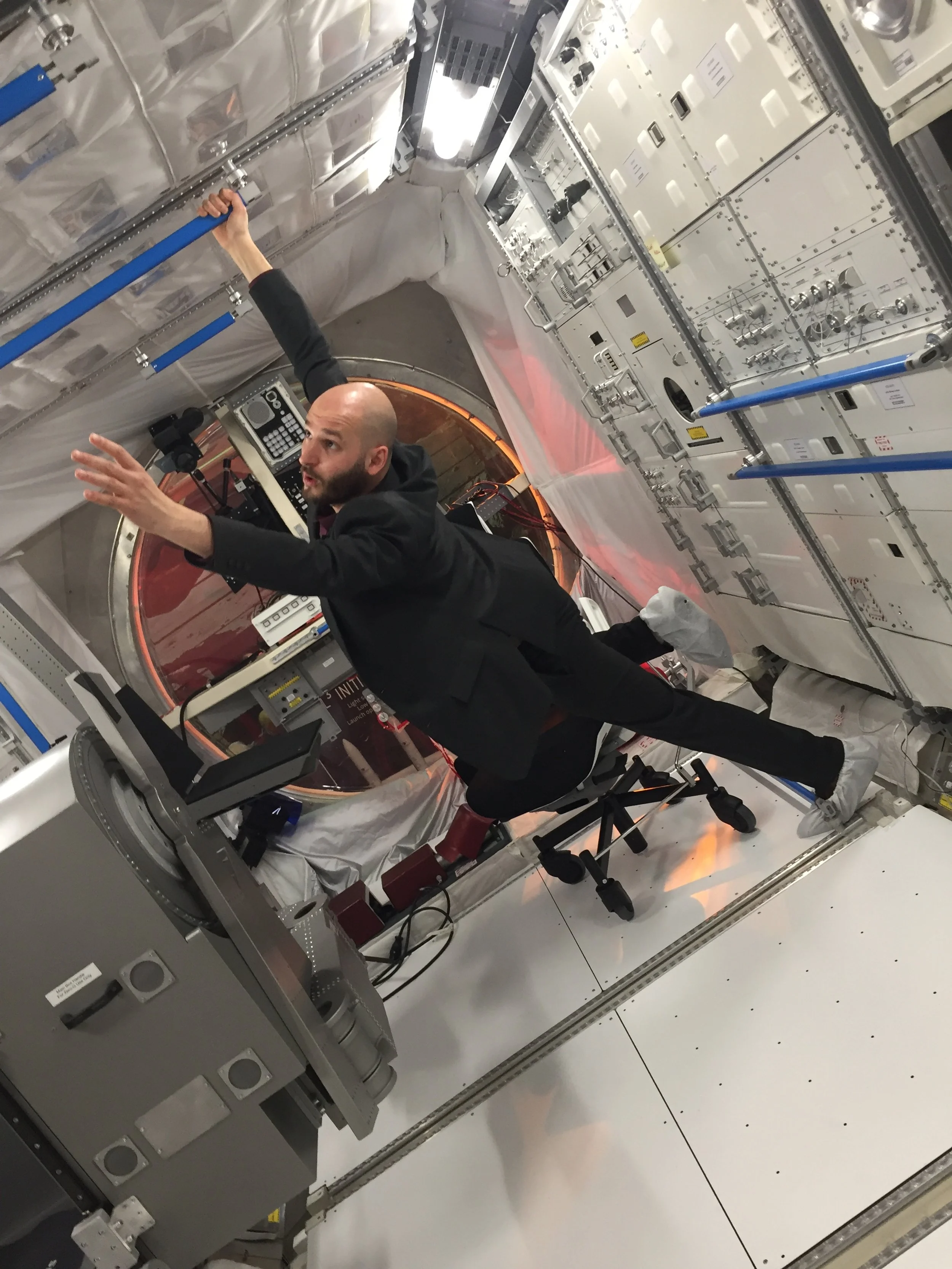 A man in a black suit is working inside a highly technical and sterile environment, possibly a laboratory or spaceship module, with complex machinery, control panels, and blue-handled tools attached to the walls.