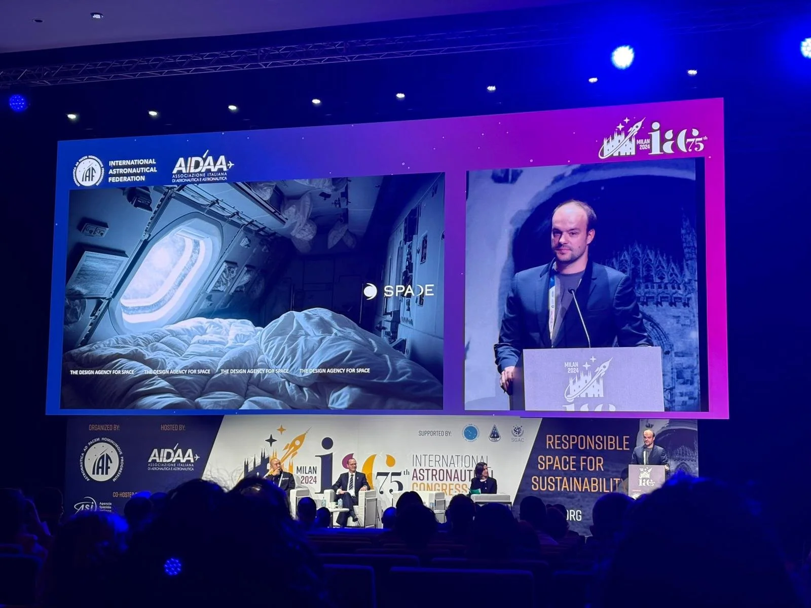 A large conference hall with a stage featuring a giant screen displaying images of a space habitat and a man speaking at a podium during the International Astronautical Congress in Milan 2024. The words 'Responsible Space for Sustainability' are visible on the screen, along with logos of various organizations.