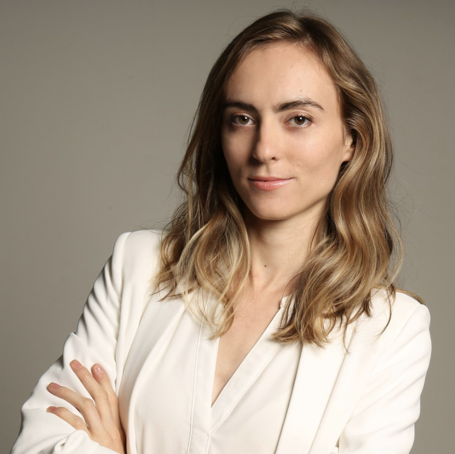 A woman with long wavy blonde hair in a white blazer, confidently looking at the camera with arms crossed against a neutral background.