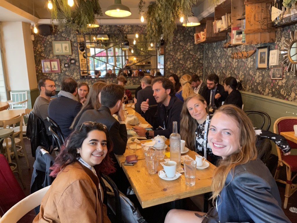 A group of young adults sitting around a long wooden table in a cozy restaurant, smiling at the camera, with drinks and coffee cups on the table, English-style decor, including framed pictures, a mirror, and hanging plants.