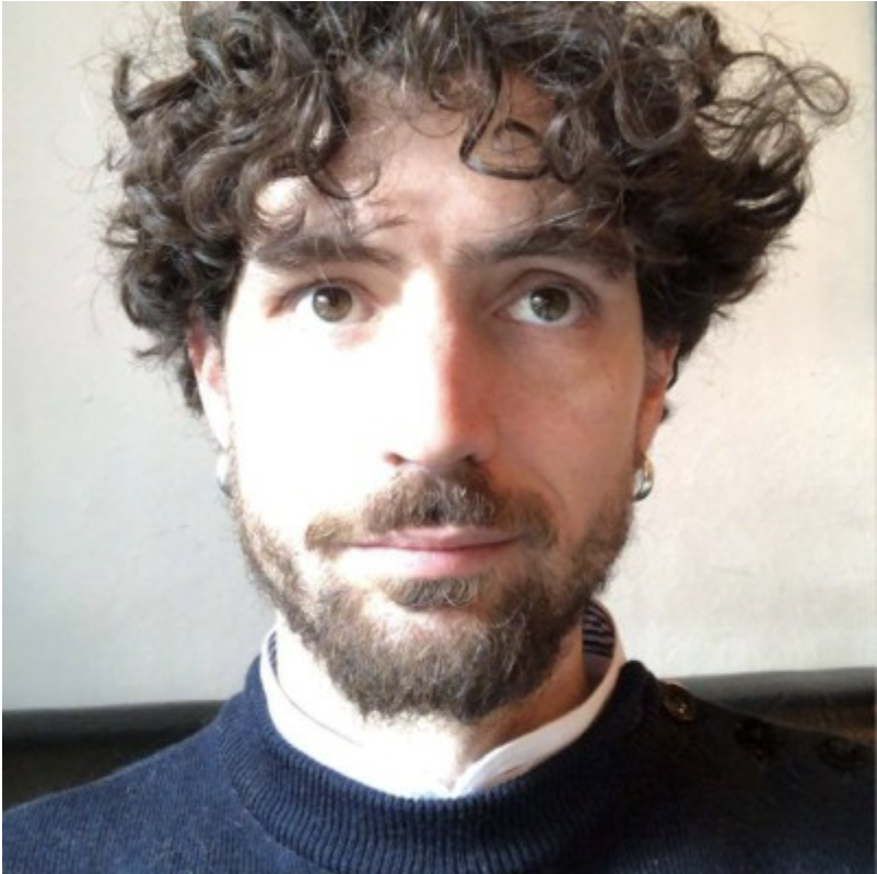 A young man with curly brown hair, light skin, a beard, and mustache, wearing a dark sweater and a striped shirt underneath, looking slightly upward in a close-up portrait.
