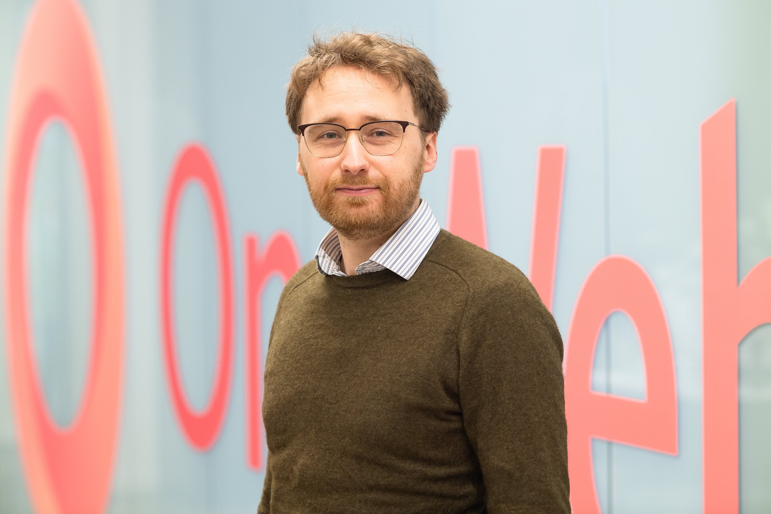 A man with glasses and a beard wearing a brown sweater over a collared shirt stands in front of a light blue wall with large pink writing.