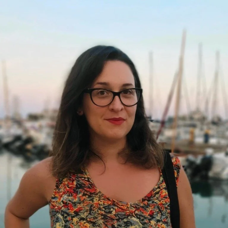 A woman with shoulder-length dark hair, wearing glasses and a colorful sleeveless top, standing in front of a marina with boats and sailboats at sunset.
