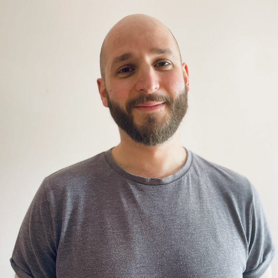 A man with a beard smiling, wearing a gray t-shirt, standing against a plain white wall.
