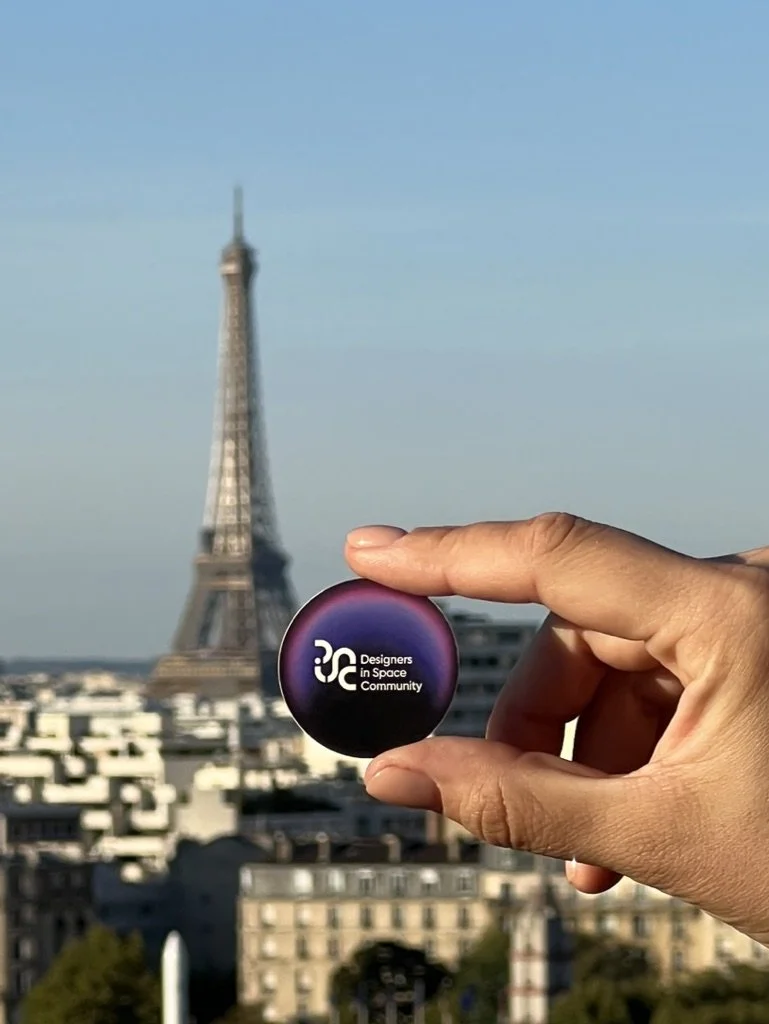 Person holding a circular badge with the logo 'Designers in Space Community' in front of the Eiffel Tower in Paris, France.