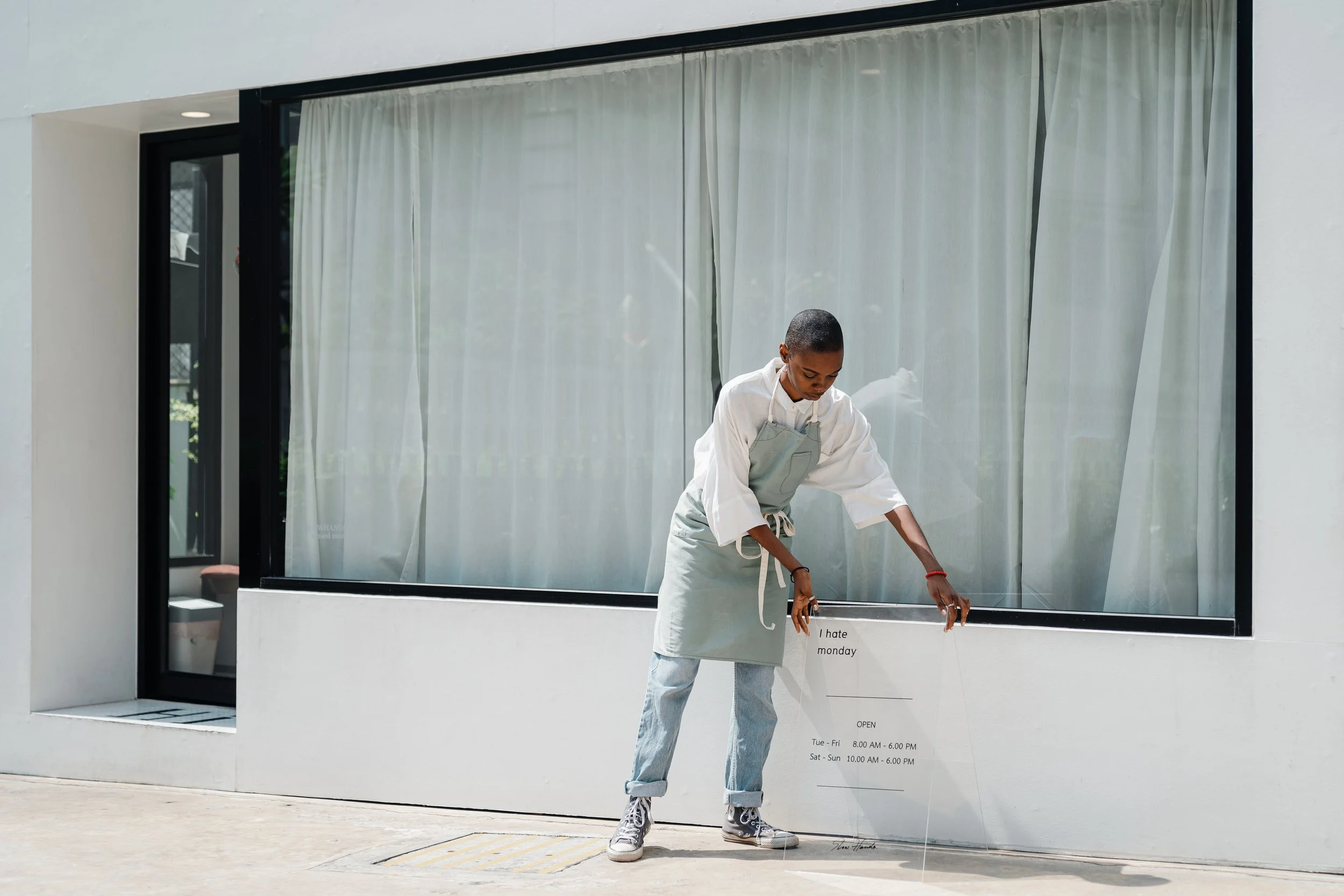 Another Good Day Maxine Hassing Impact Businesses Grow and Build Your Business. We are a Strategy and Marketing Agency Entrepreneurs. person dressed as a chef adjusting a restaurant sign outside a modern building with large window and white curtains.