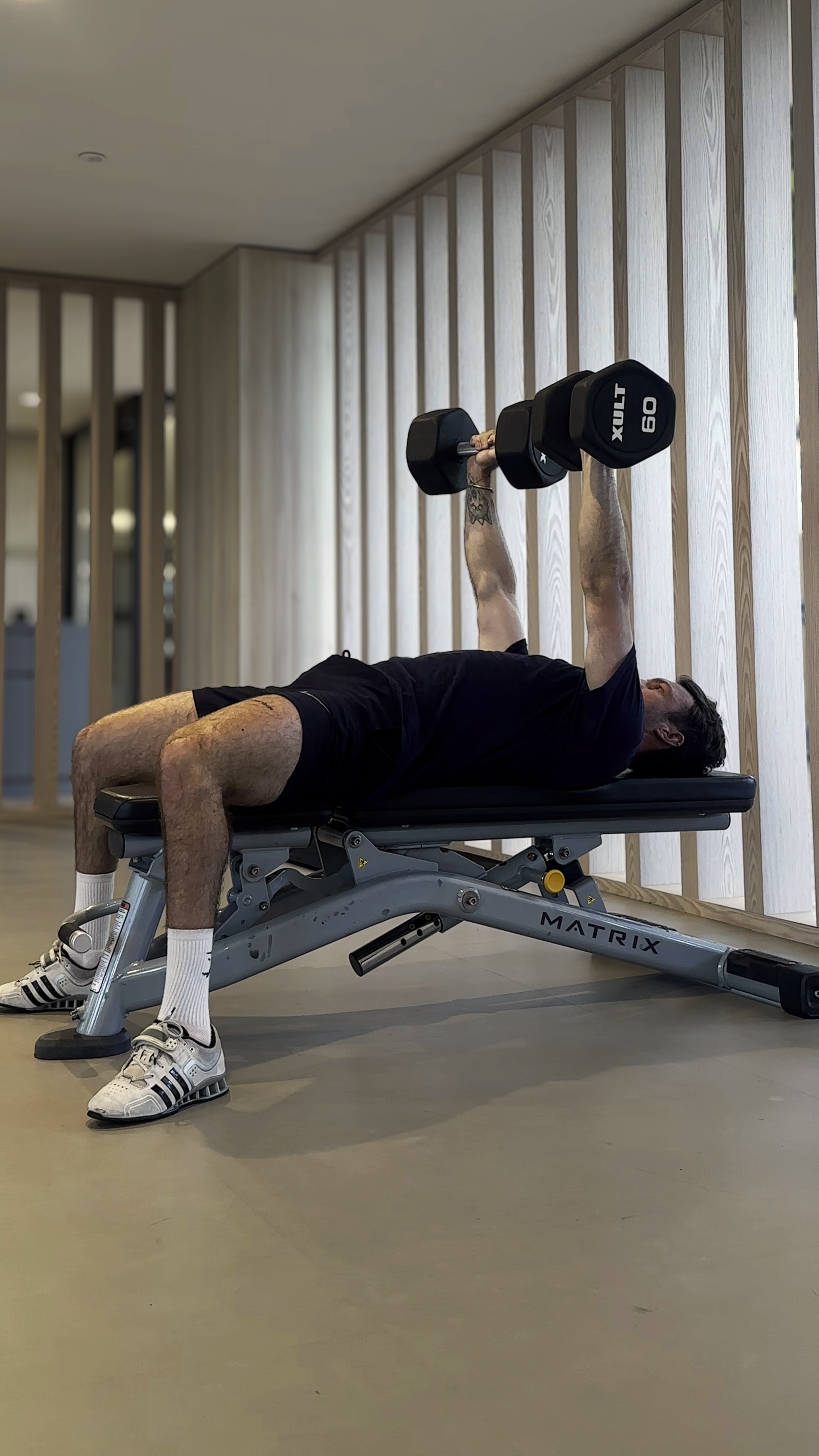 A man lying on a gym bench doing a dumbbell chest press exercise, wearing black shorts, a black t-shirt, white socks, and sneakers, in a modern fitness facility with wooden wall panels.