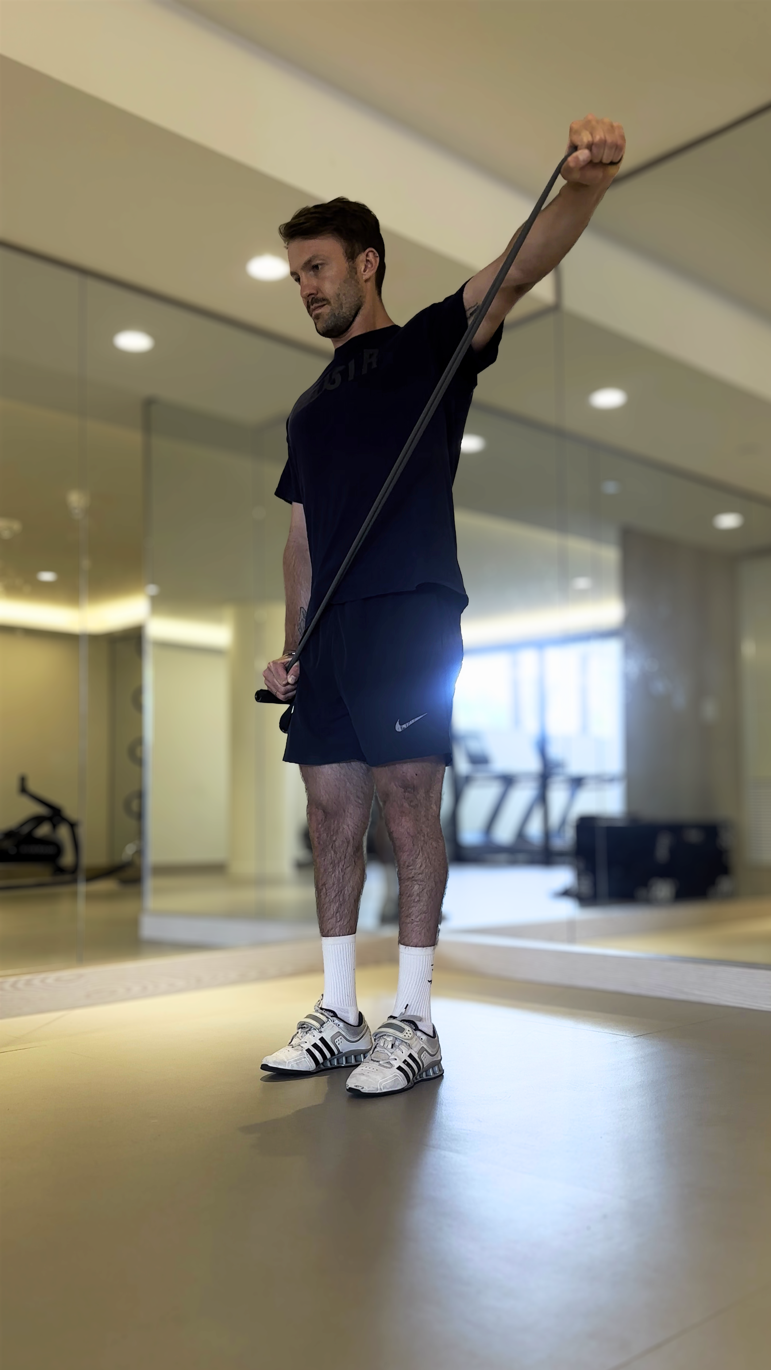 Man in a gym performing a stretching exercise with a resistance band, wearing athletic shorts, a t-shirt, and sneakers.