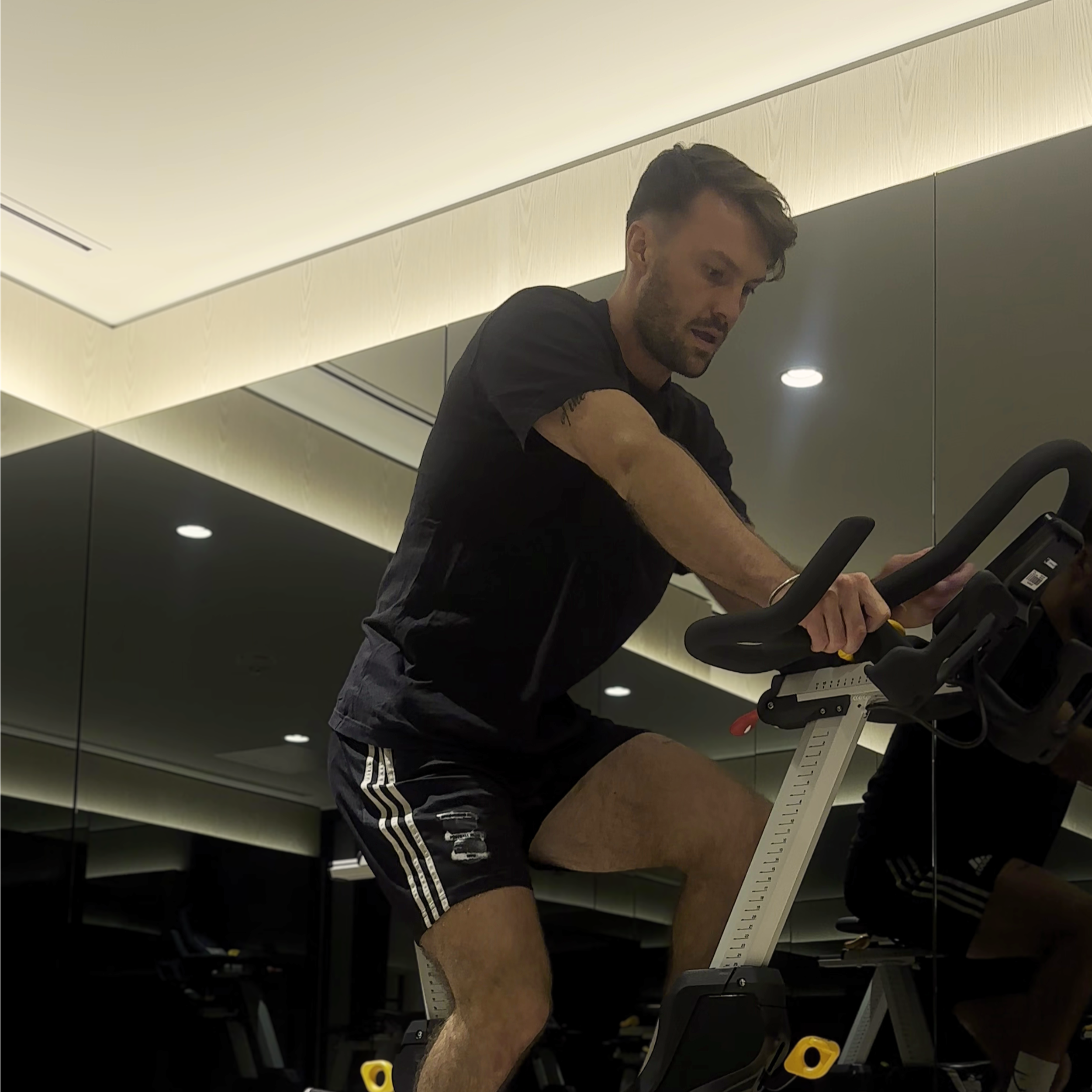 A man with dark hair and a beard, wearing a black t-shirt, is exercising on a stationary bike in a gym. The gym has light-colored walls with a mirror reflecting the ceiling lights.