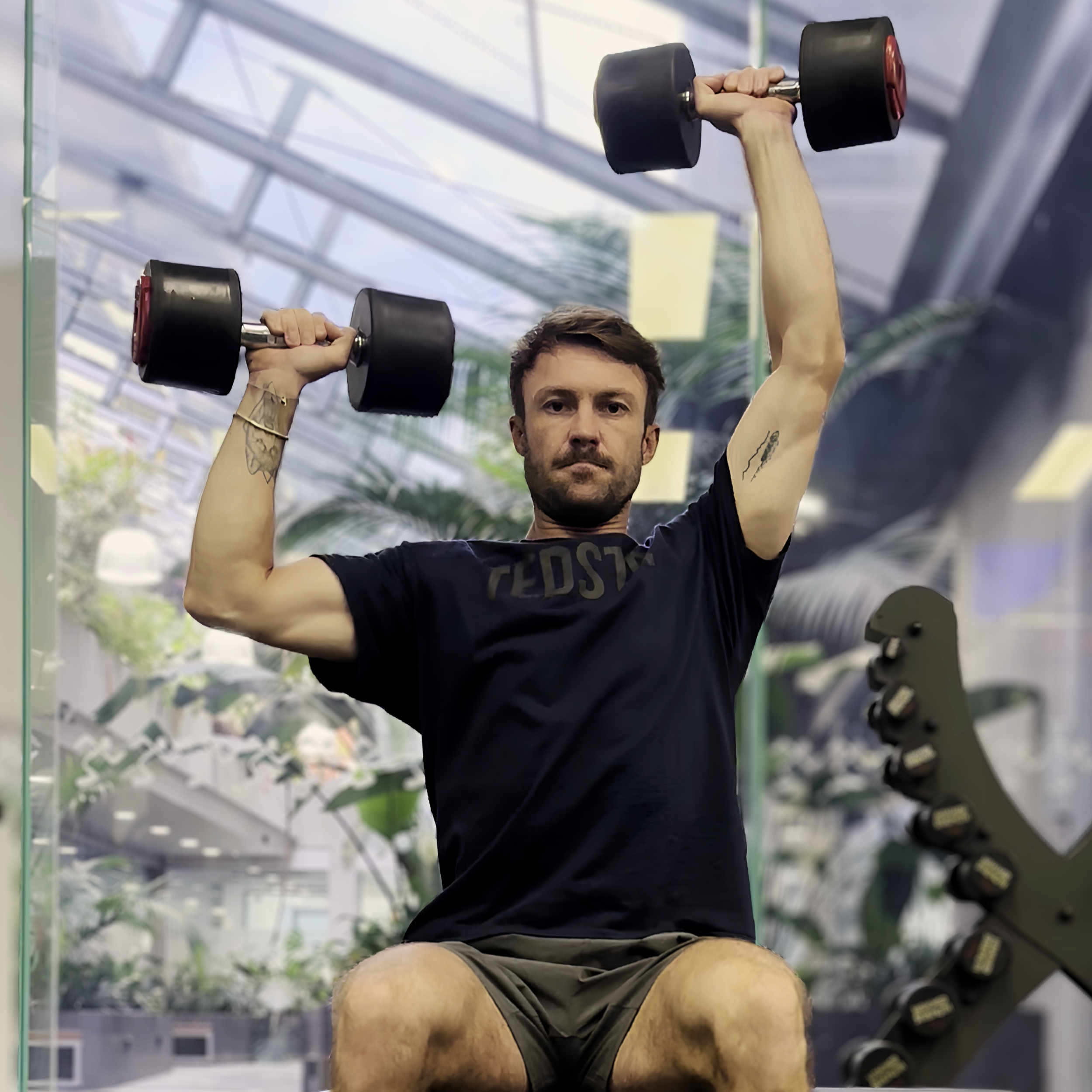 A man lifting a black dumbbell in a modern gym with glass walls and visible ceiling structure