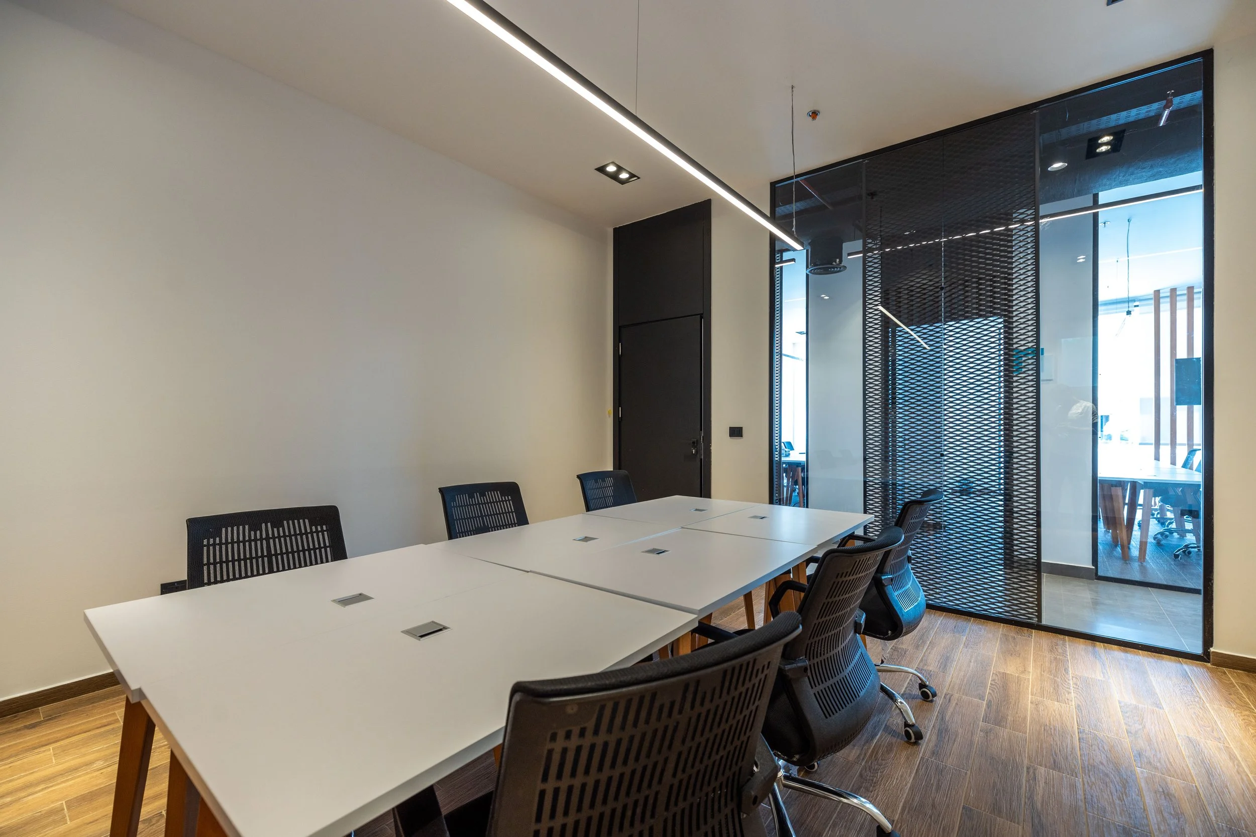 Empty modern conference room with black chairs around a white table, glass wall with black frame, and wood flooring.