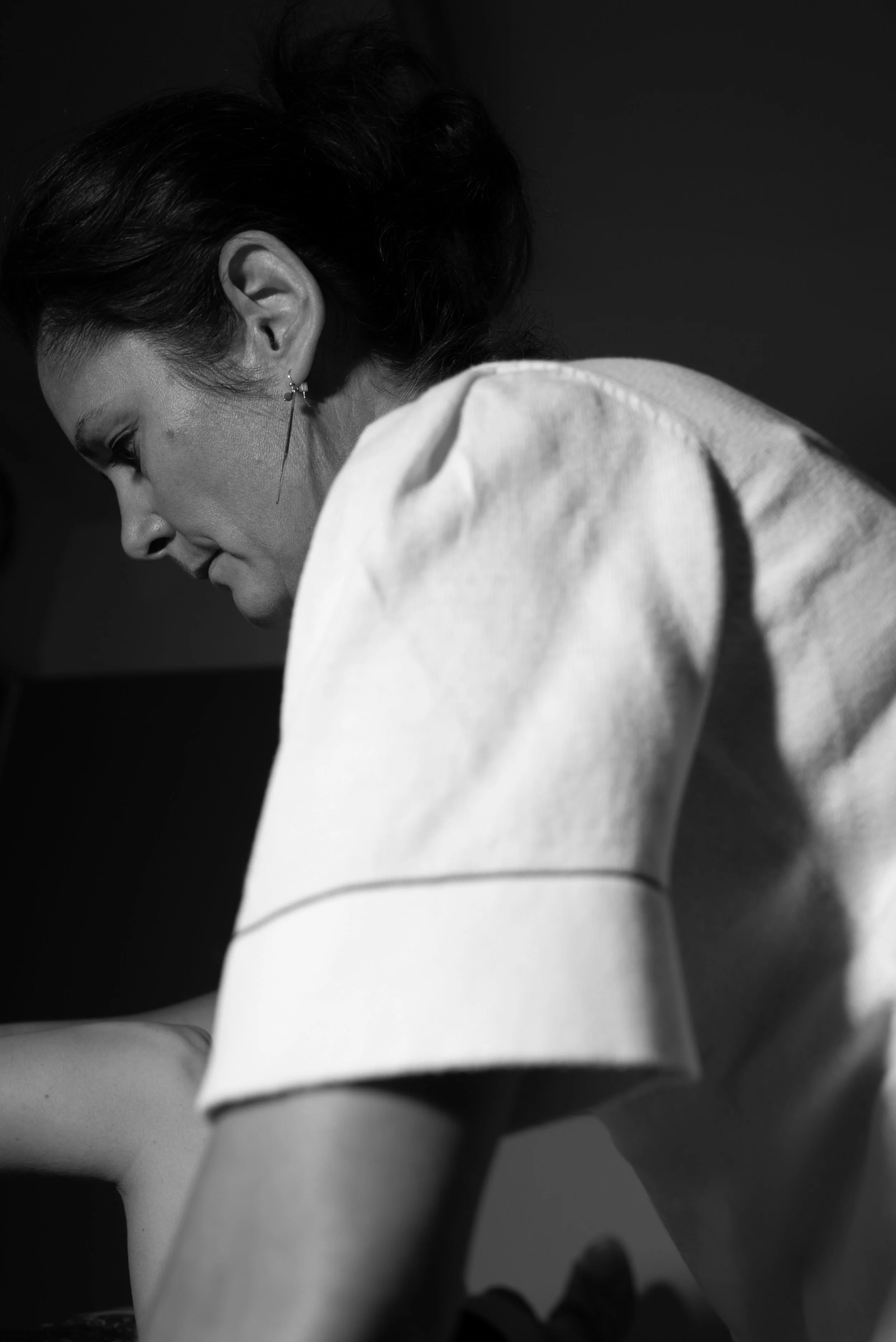Zwarte en witte close-up van een vrouw met donker haar, gekleed in een witte blouse, die naar beneden kijkt.