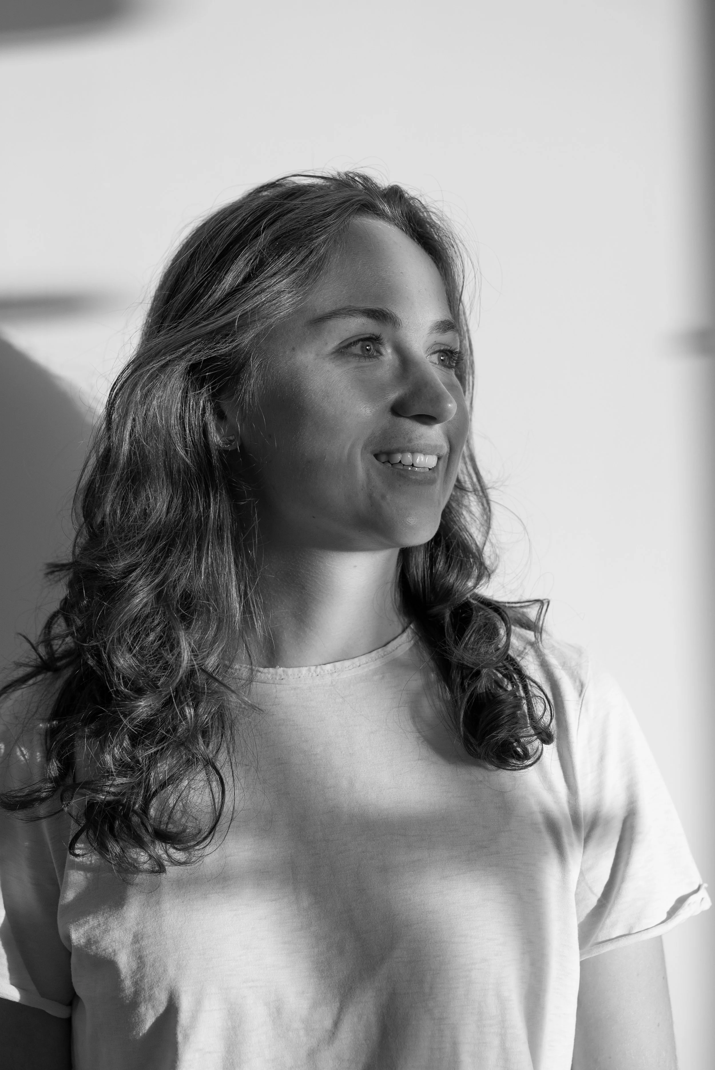 Portret van een jonge vrouw met bruin, golvend haar die glimlacht, gekleed in een lichte T-shirt, tijkend in een ruimte met lichte muren, in zwart-wit fotografie.