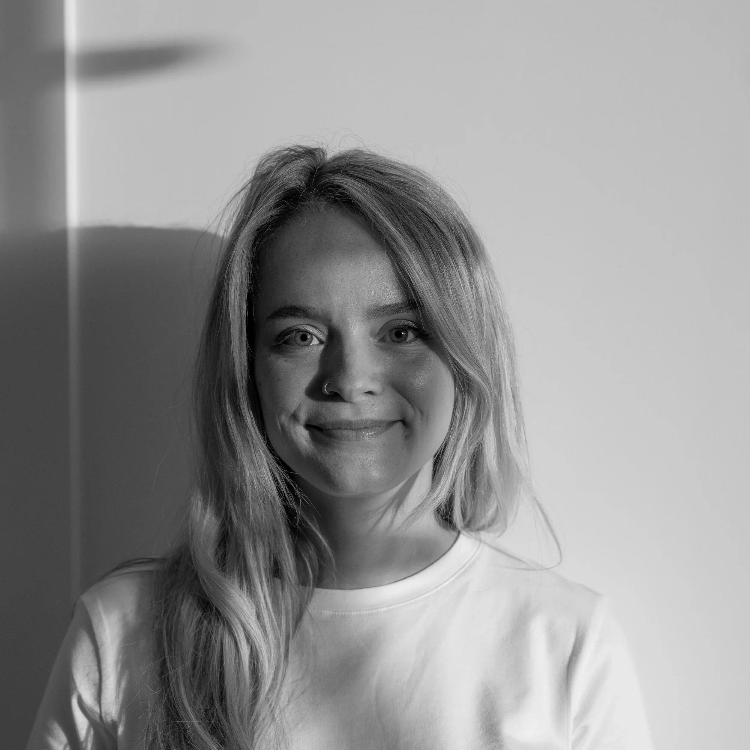 Zwart-wit foto van een glimlachende jonge vrouw met lang haar en een neuspiercing, zittend voor een lichte achtergrond.
