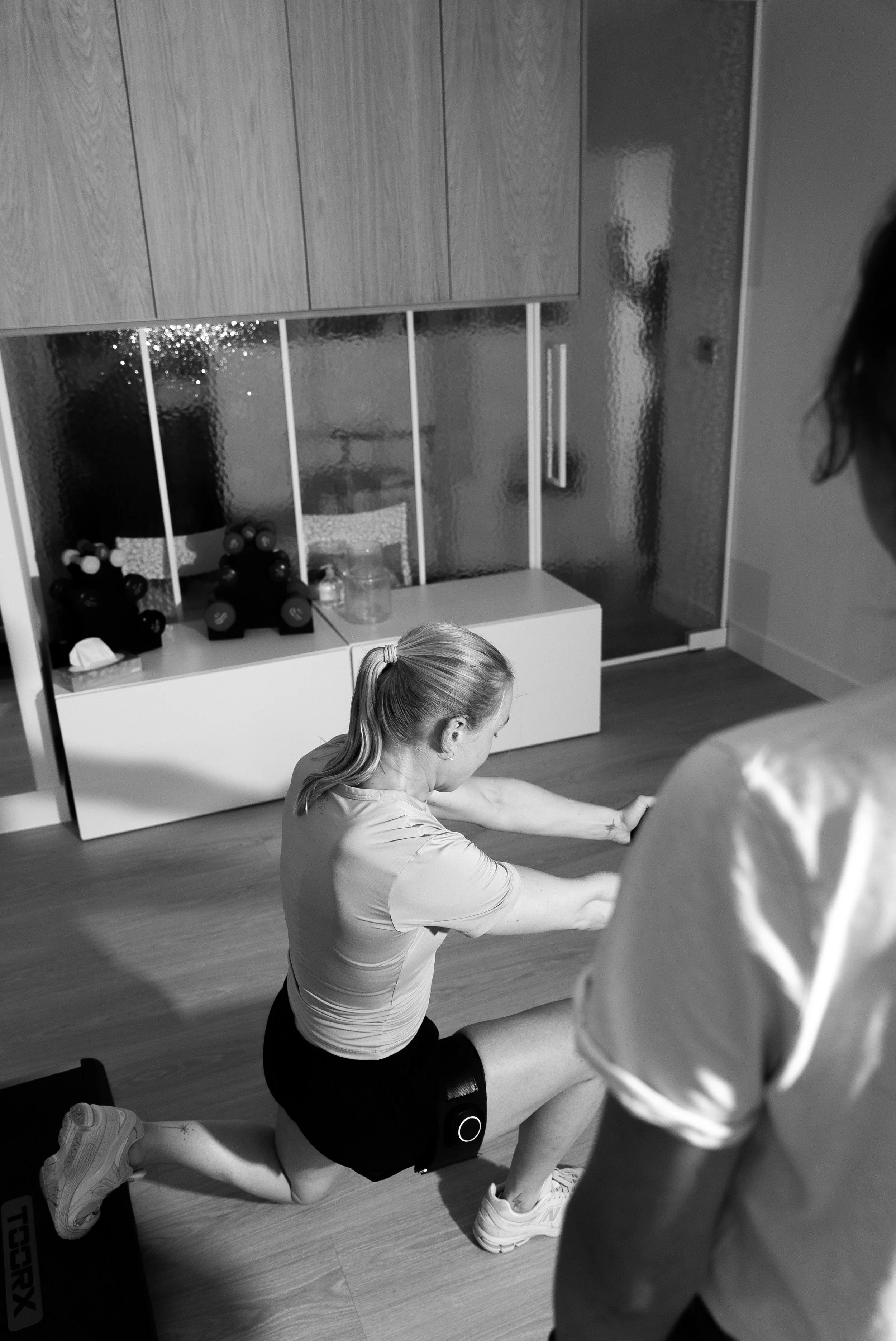 Een vrouw doet een lunges-oefening terwijl een persoon haar observeert in een binnenruimte.