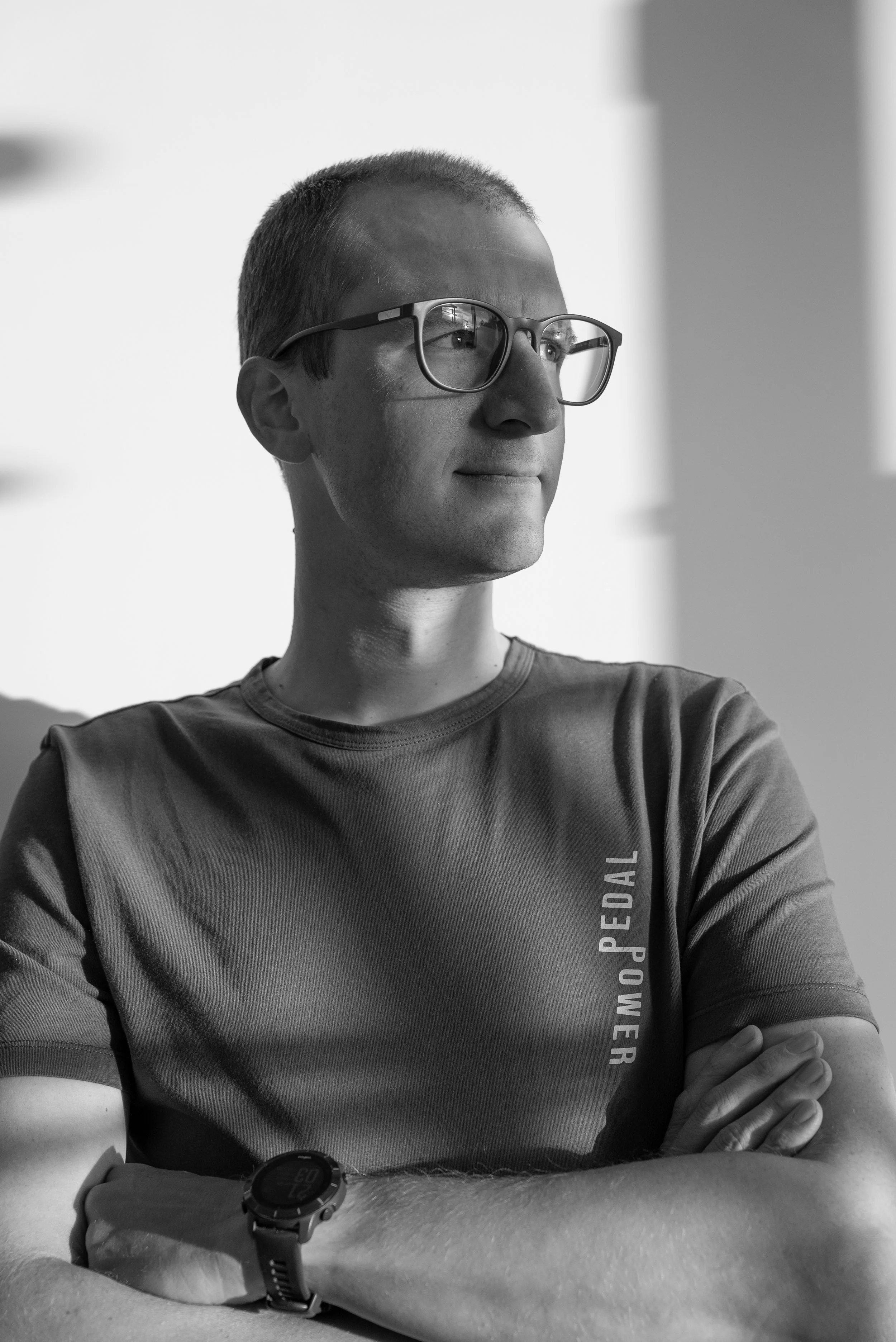Zwart-wit foto van een man met korte haar, bril en een T-shirt met de tekst 'POWDER' die in de schaduw en het licht kijkt met zijn armen over elkaar.