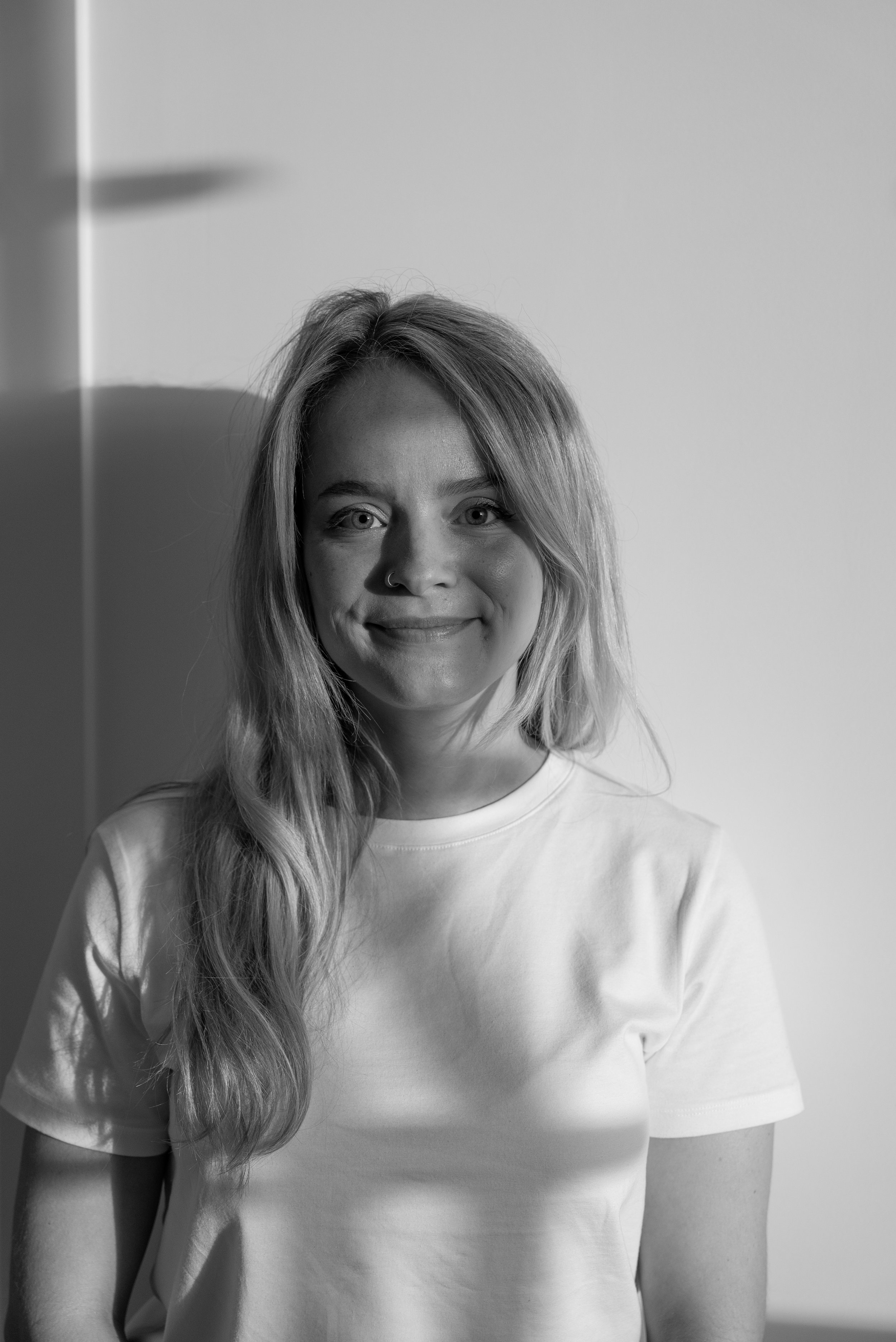 Een portretfoto van een glimlachende vrouw met lang, golvend haar, gekleed in een wit T-shirt, op een neutrale achtergrond.